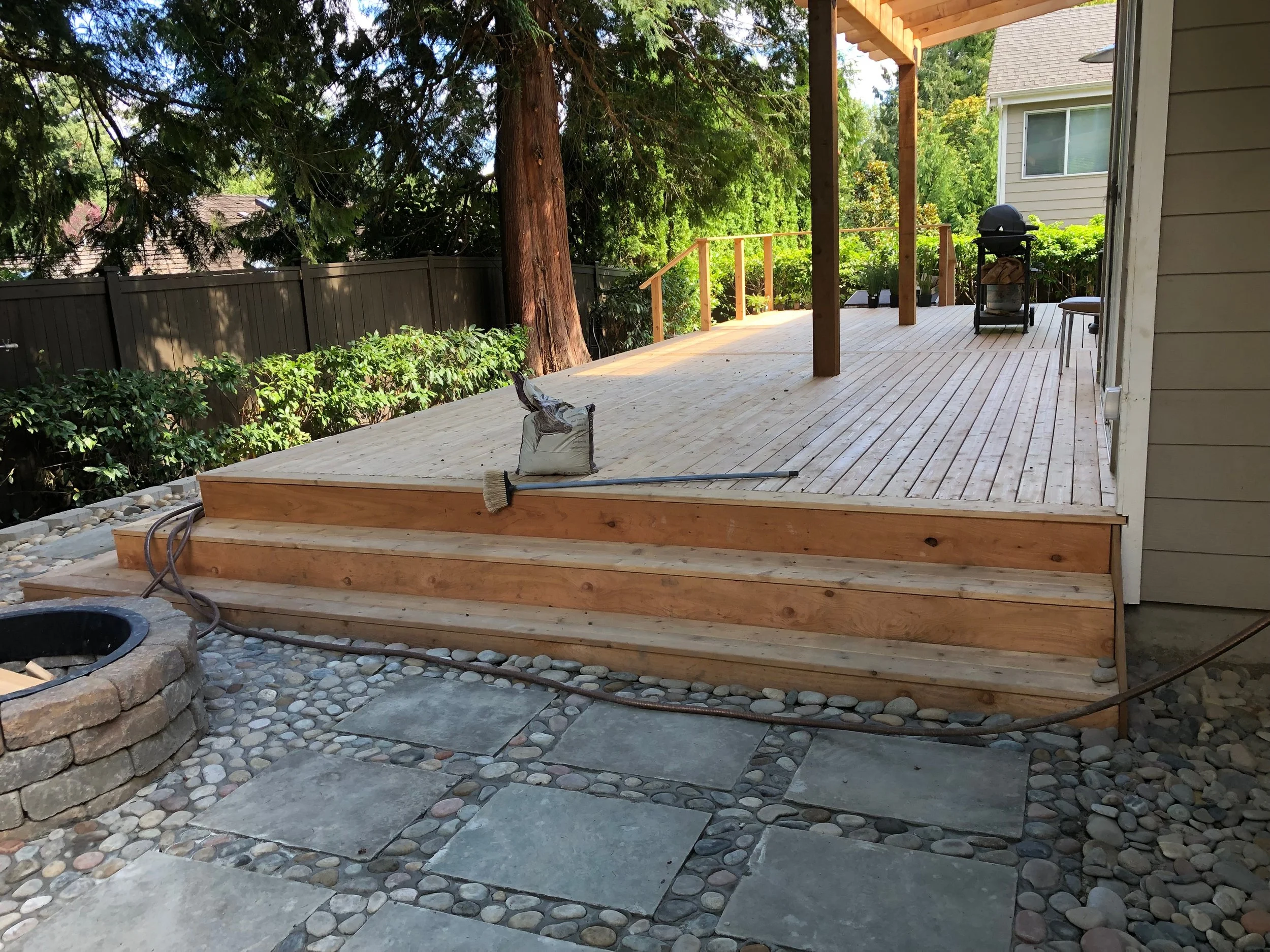 Backyard patio with wooden deck, steps, a BBQ grill, a chair, lush trees, and a stone and pebble garden area.