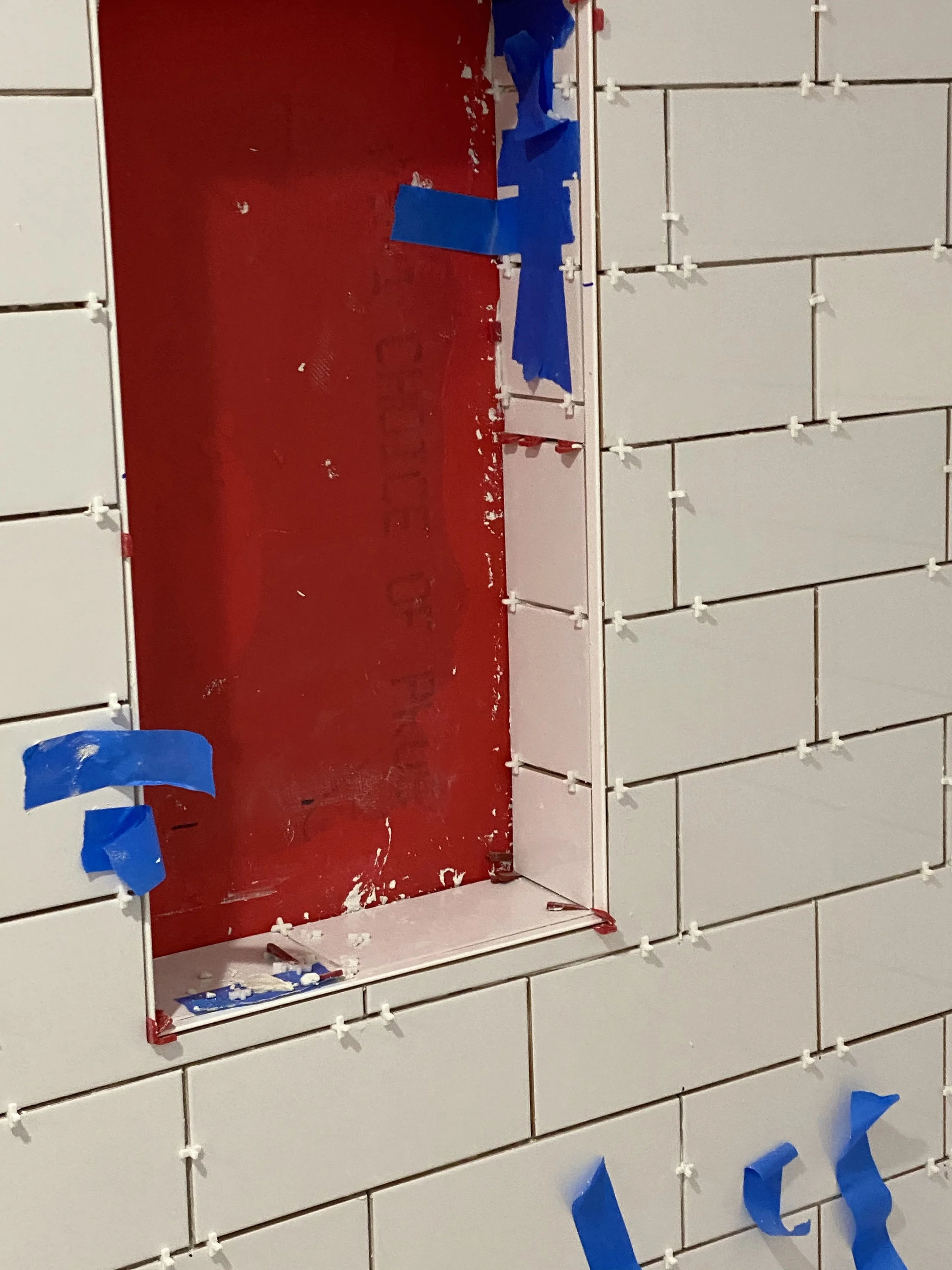 Close-up view of wall tiles with a red background and white tiles, some blue painter's tape on the tiles, and white plastic spacers in between tiles, indicating a tile installation process.