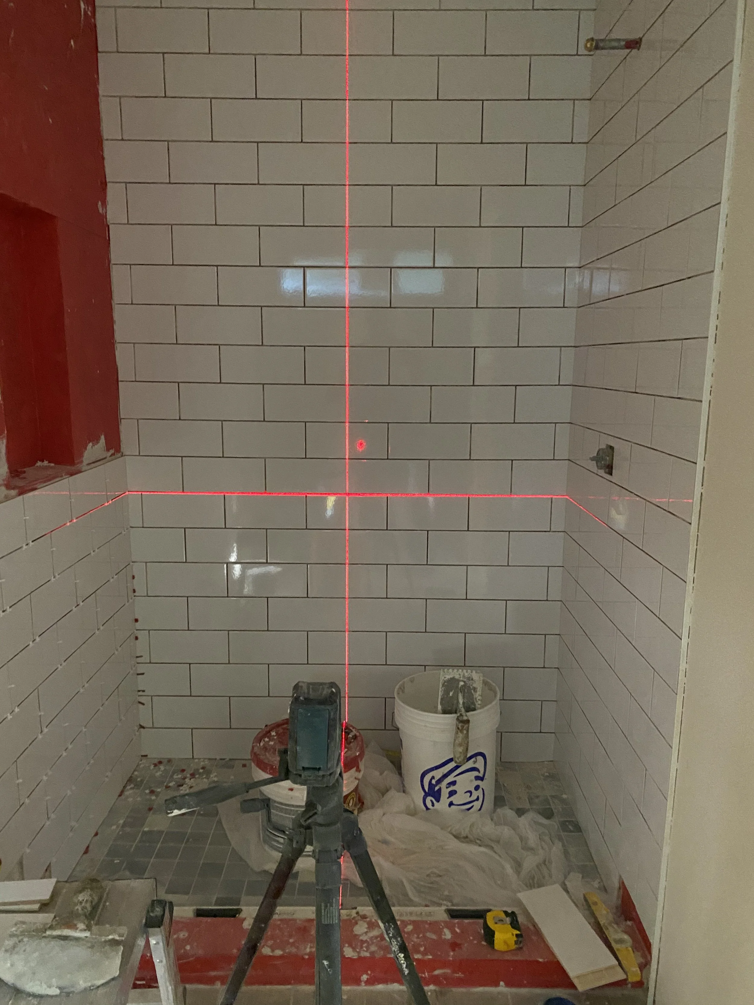 A bathroom under construction with white subway tiles, a red wall on the left, and construction tools and supplies, including a laser level projecting a red line, a bucket, and a saw, on the floor.