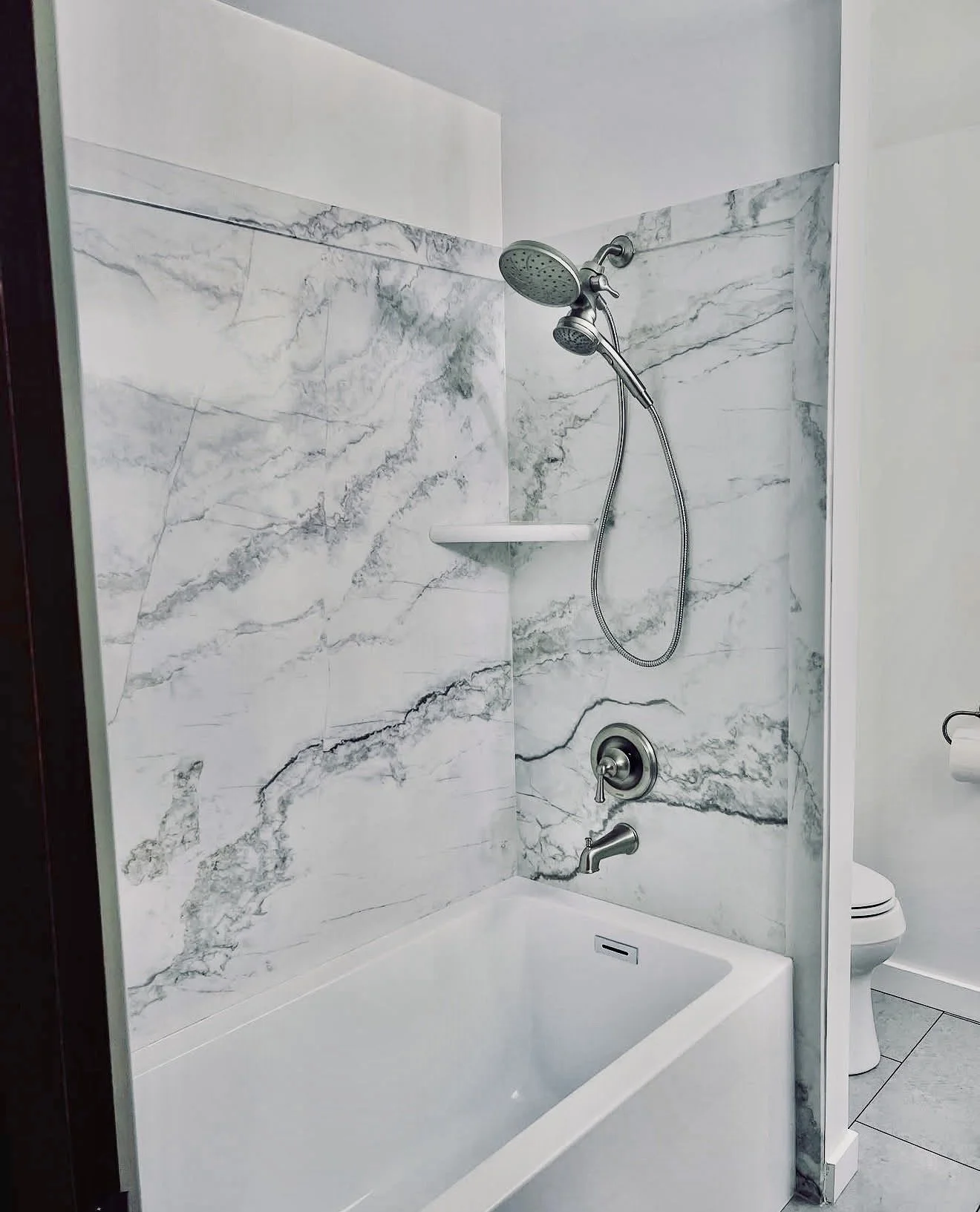 A modern shower area with marble walls featuring prominent gray veining, a handheld showerhead, and a bathtub with a drain cover. Adjacent to the shower area, there's a toilet visible in the background.