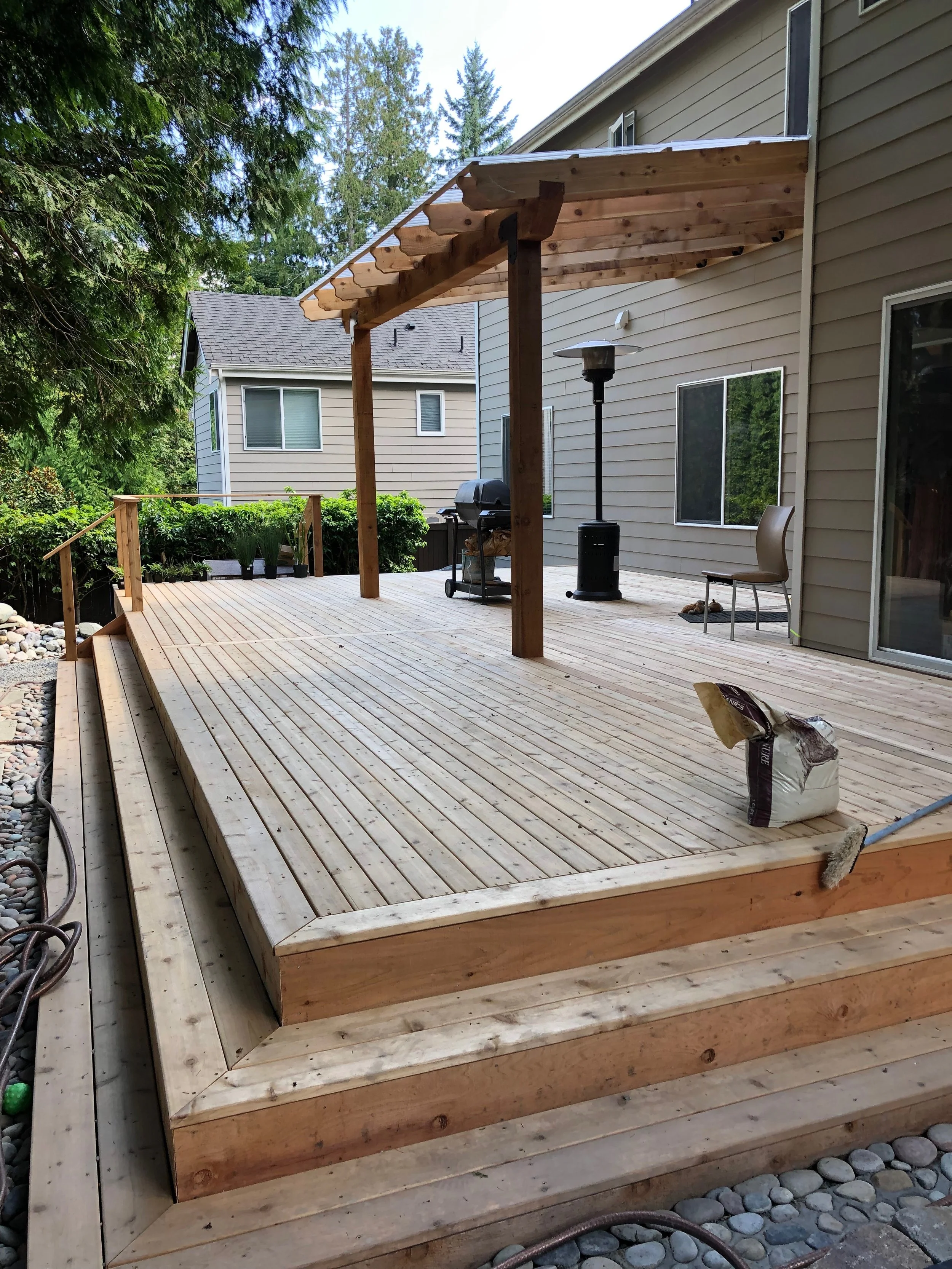 View of a wooden deck under construction, with stairs leading up to the deck, a chair, a patio heater, and some gardening plants in the background.