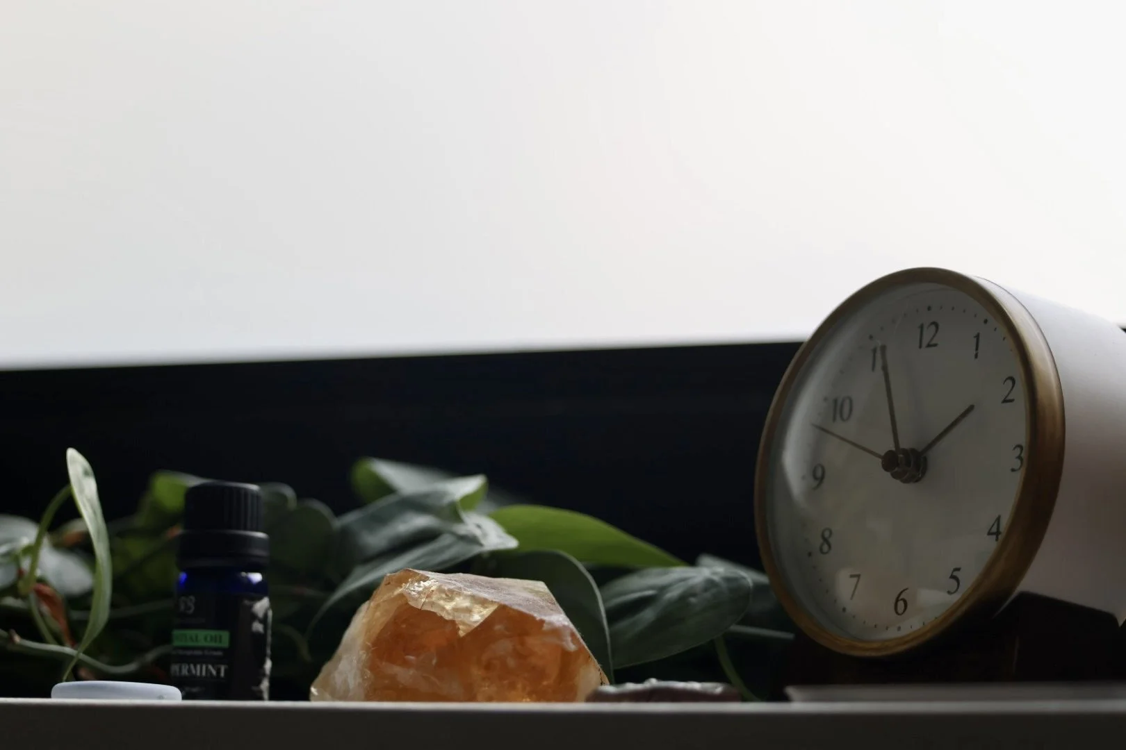 A white and gold alarm clock showing 2:55, a small bottle of mint essential oil, a large orange crystal, and green foliage on a surface with a window in the background.
