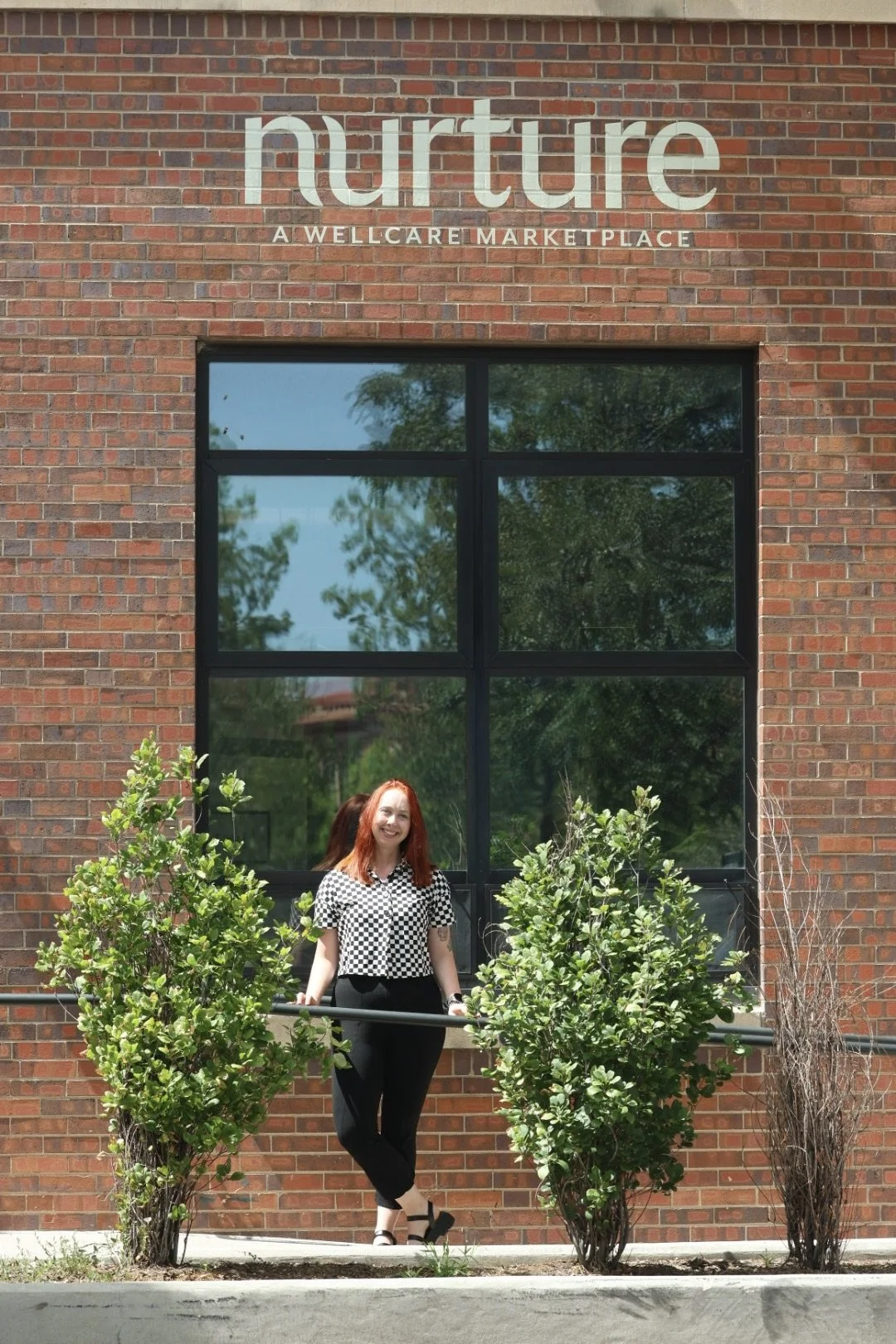 A woman with red hair wearing a black and white checkered shirt, standing outside in front of a brick building with large window and a sign reading 'nurture: A Wellcare Marketplace', surrounded by green bushes.