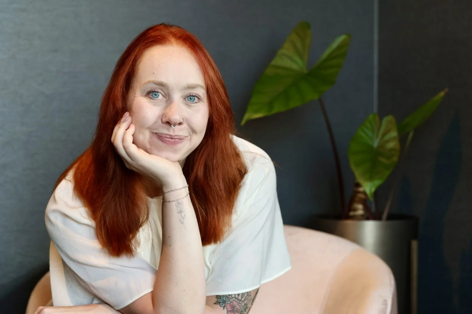 A woman with red hair, blue eyes, and a septum piercing, smiling and resting her chin on her hand, sitting in a room with a dark green wall and a potted plant with large green leaves in the background.