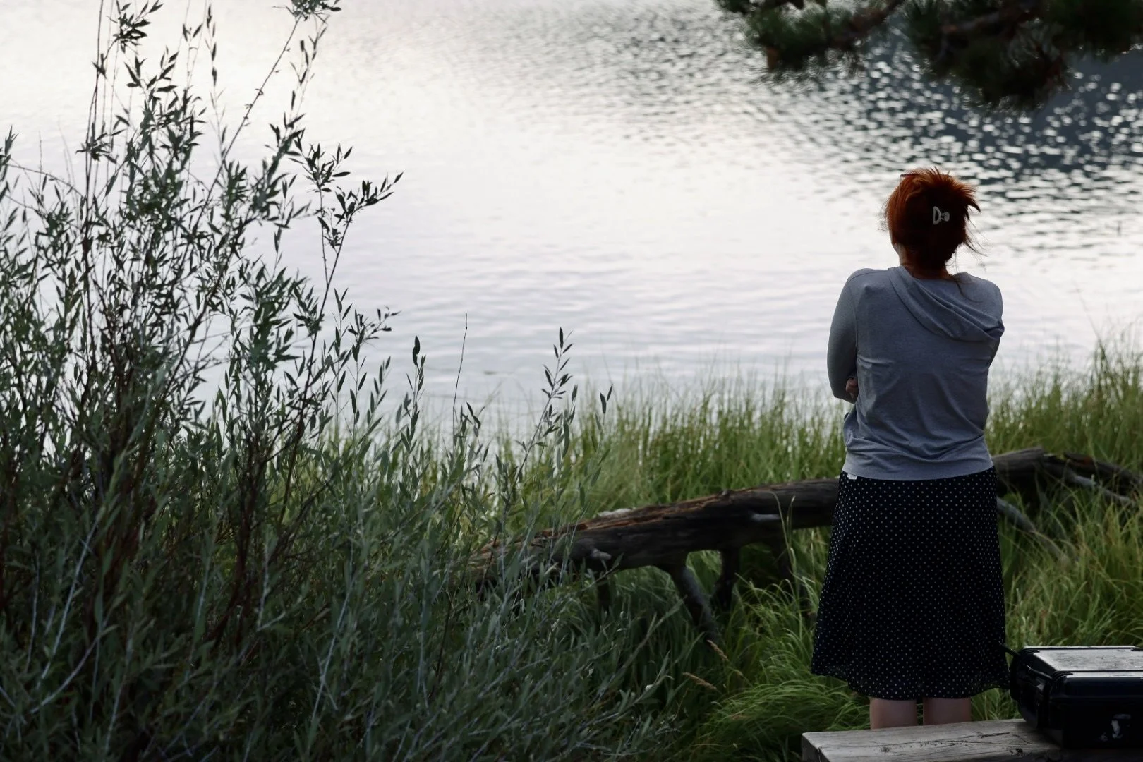A woman with red hair, wearing a gray hoodie and black polka dot skirt, stands on a wooden deck near a lake, looking at the water surrounded by tall green grass and bushes.