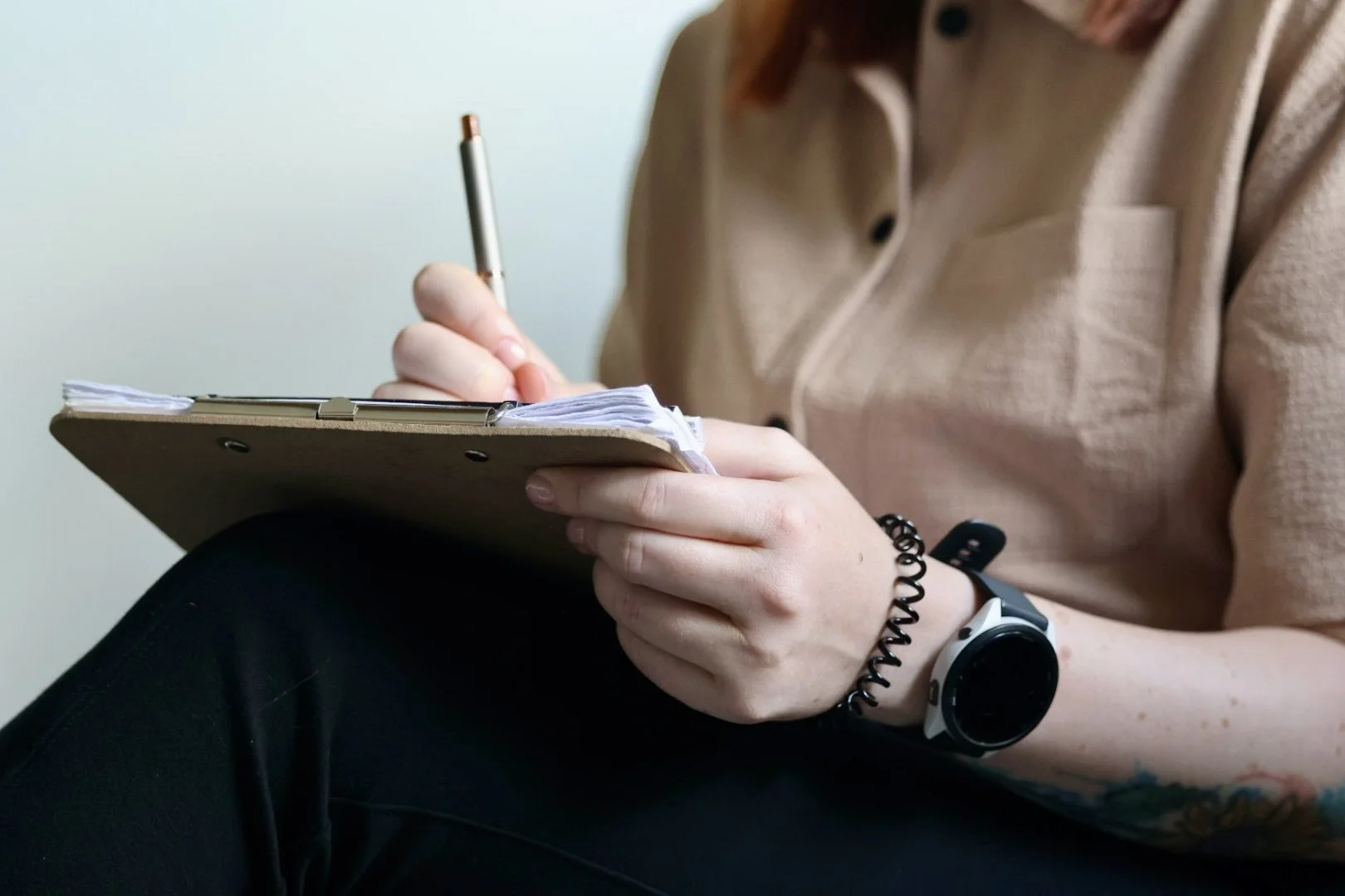 A person sitting and taking notes in a small notebook with a pen, wearing a beige shirt and black pants, with a smartwatch on their wrist.