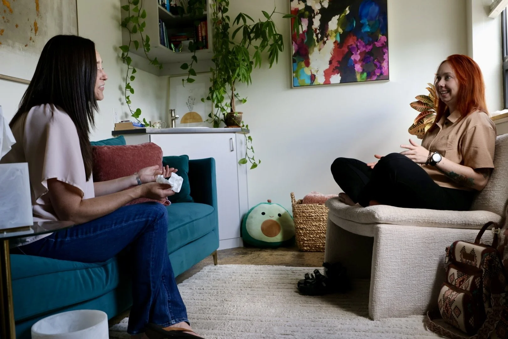 Two women sitting and talking in a cozy living room with plants, artwork, and a plush avocado-shaped cushion.