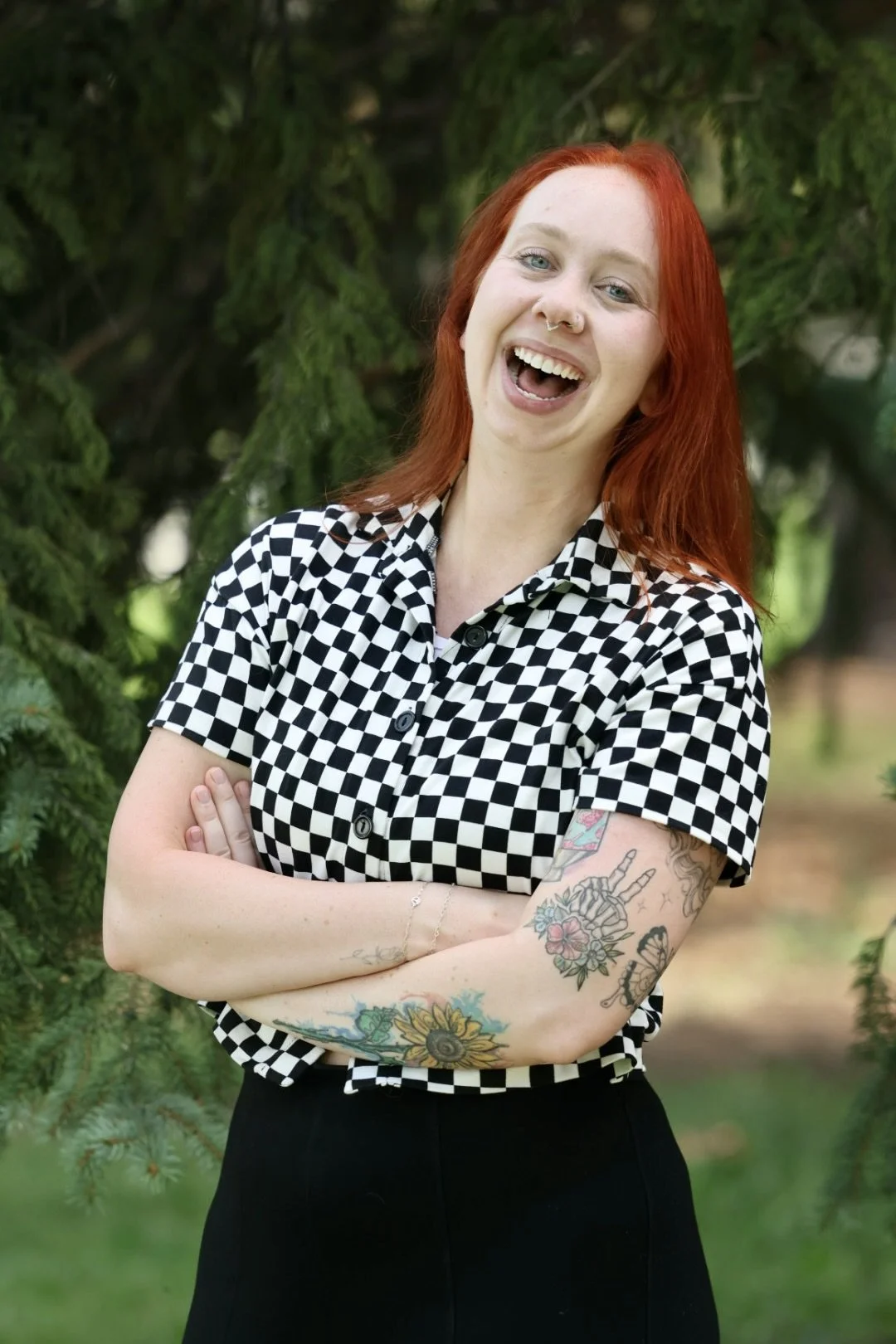 A young woman with red hair, tattoos on her arms, wearing a black and white checkered shirt, smiling with her arms crossed outdoors near green trees.