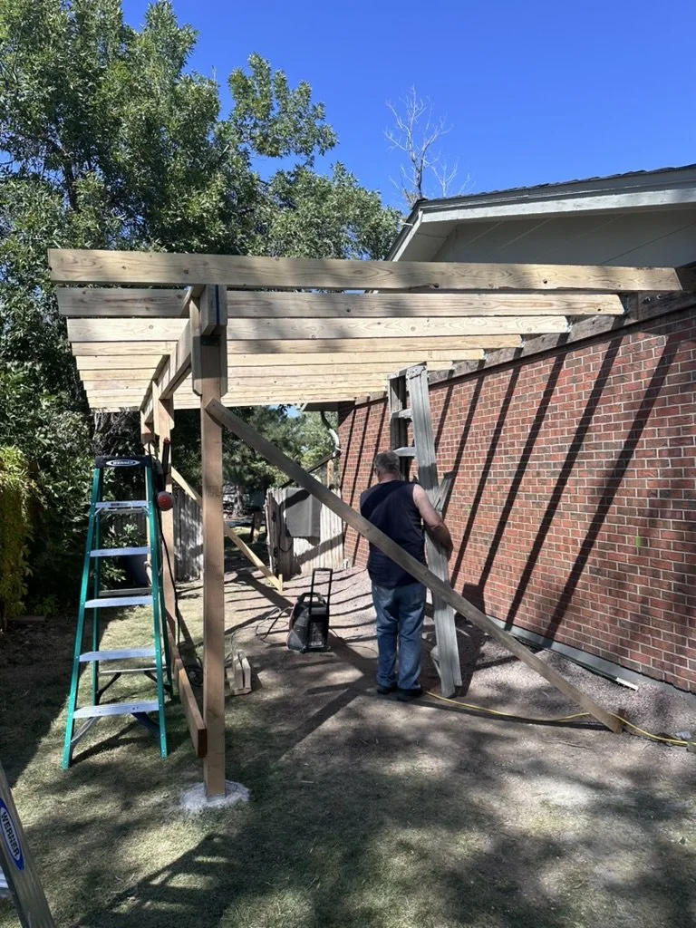 A contractor building a car port or extension attached to a brick garage.  There is a ladder leaning against the structure, tools and materials around, and shadows cast by the wooden beams.