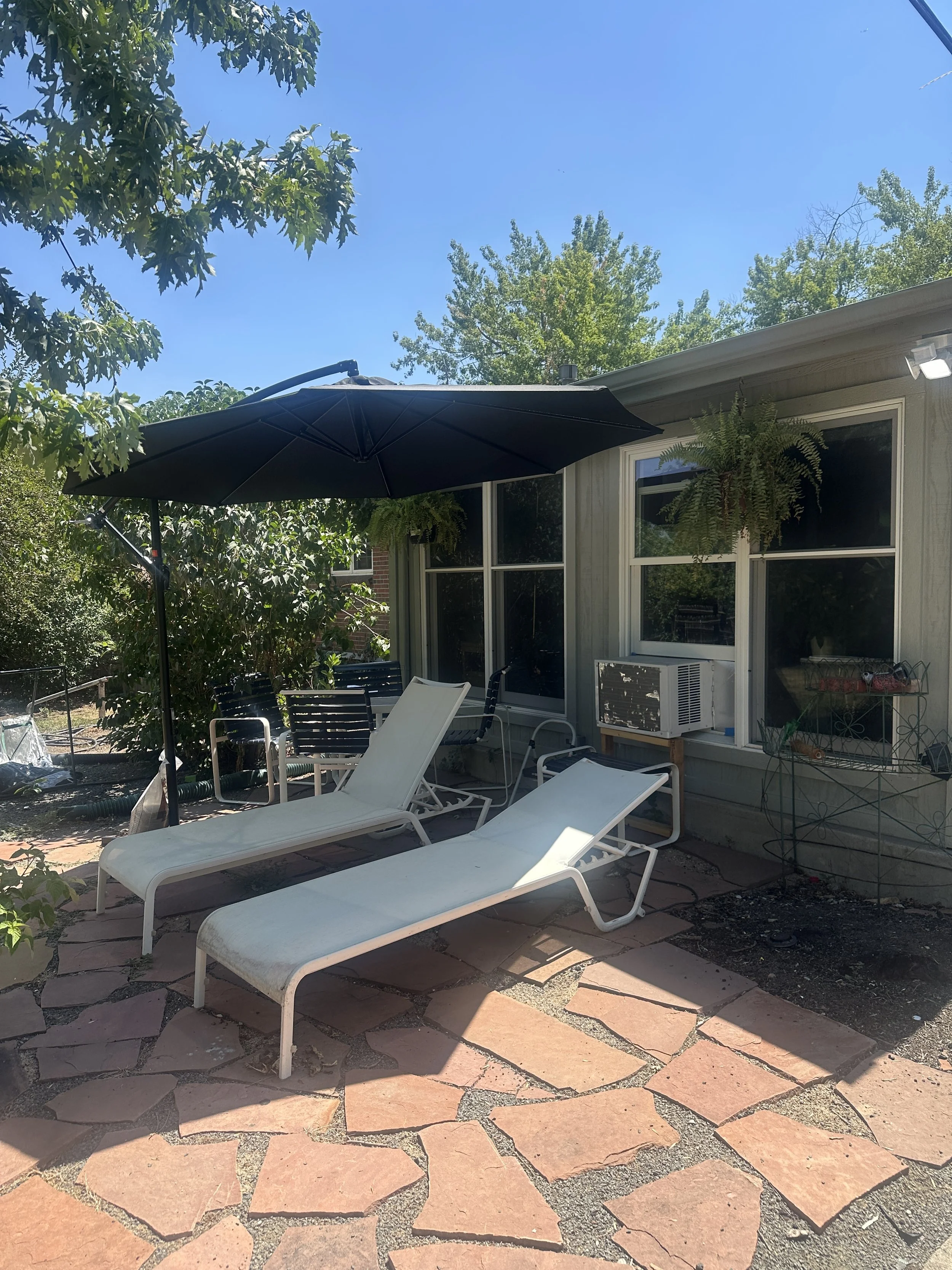 Backyard patio with lounge chairs, an umbrella, potted plants, and a house wall with windows.