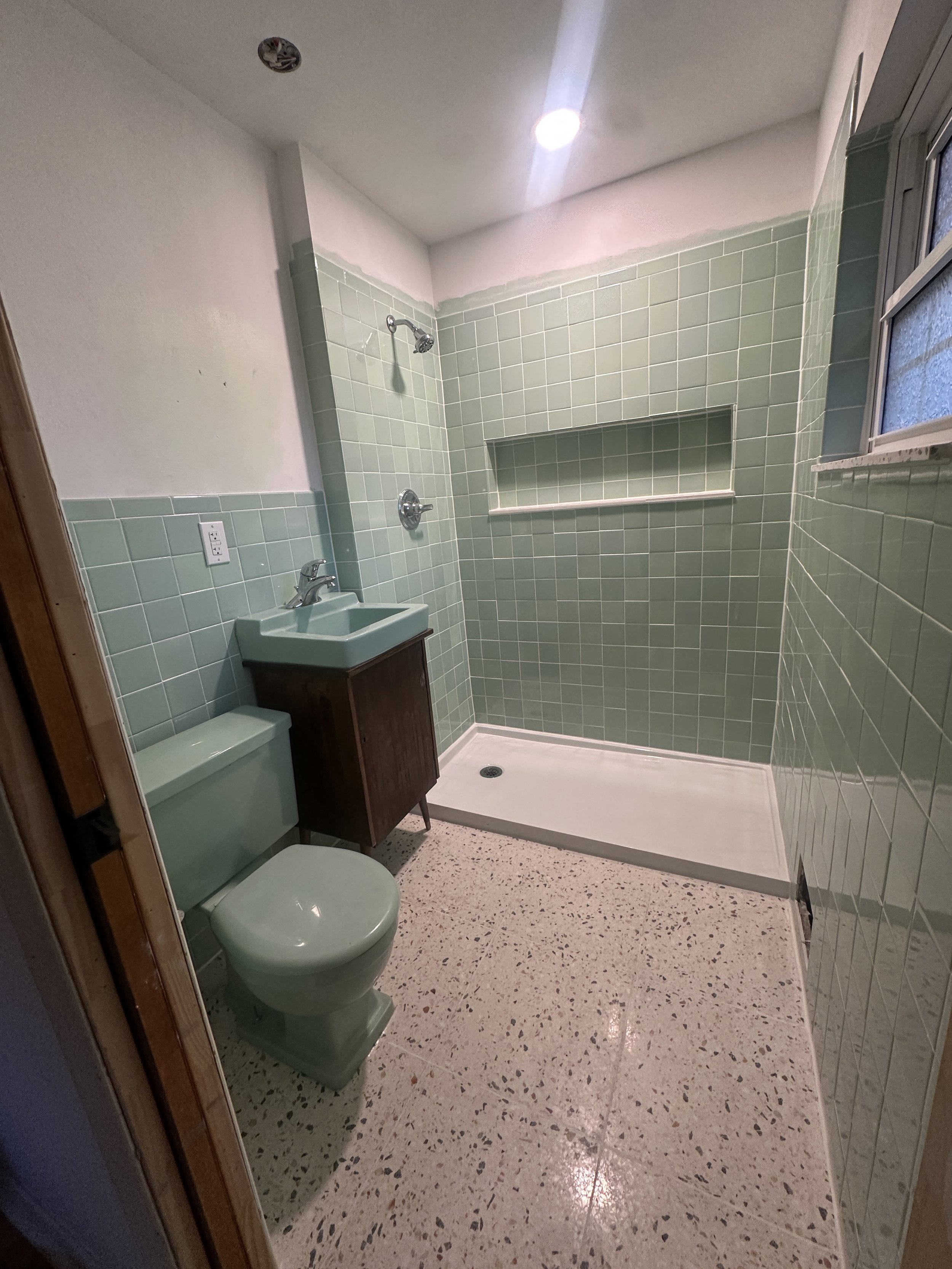 A bathroom with green tiled walls, a green toilet, a small green sink with a wooden cabinet, a shower area with a built-in shelf, a window, and a speckled terrazzo floor.