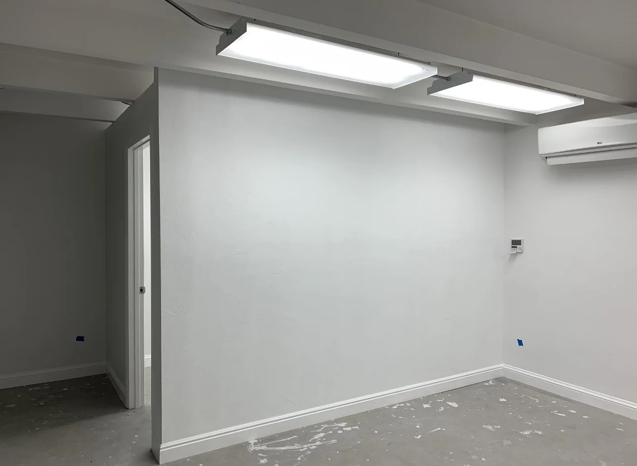 Empty room with white walls, fluorescent ceiling lights, and an air conditioning unit, with some paint splatters on the floor.