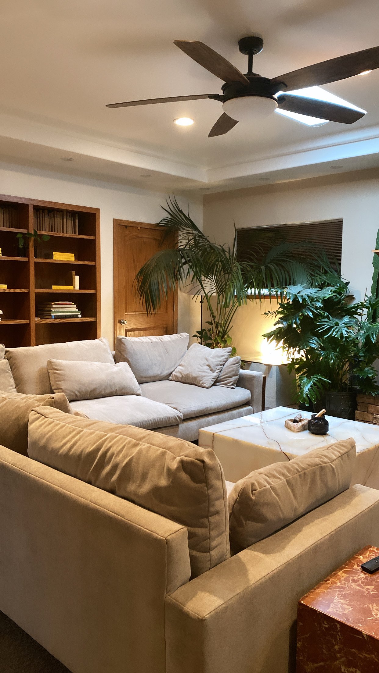 Living room with beige sofas, a marble coffee table, bookcase, ceiling fan, green plants, and a window with blinds.