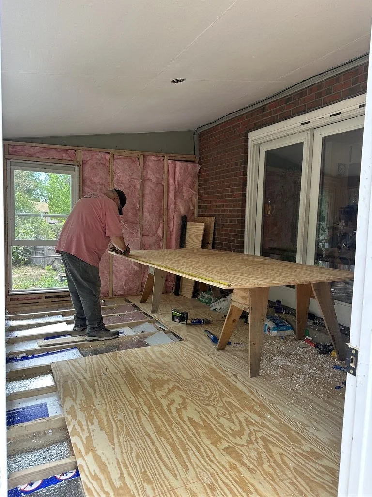 David installing a new floor inside a room with a large wooden table, pink insulation, and construction tools.