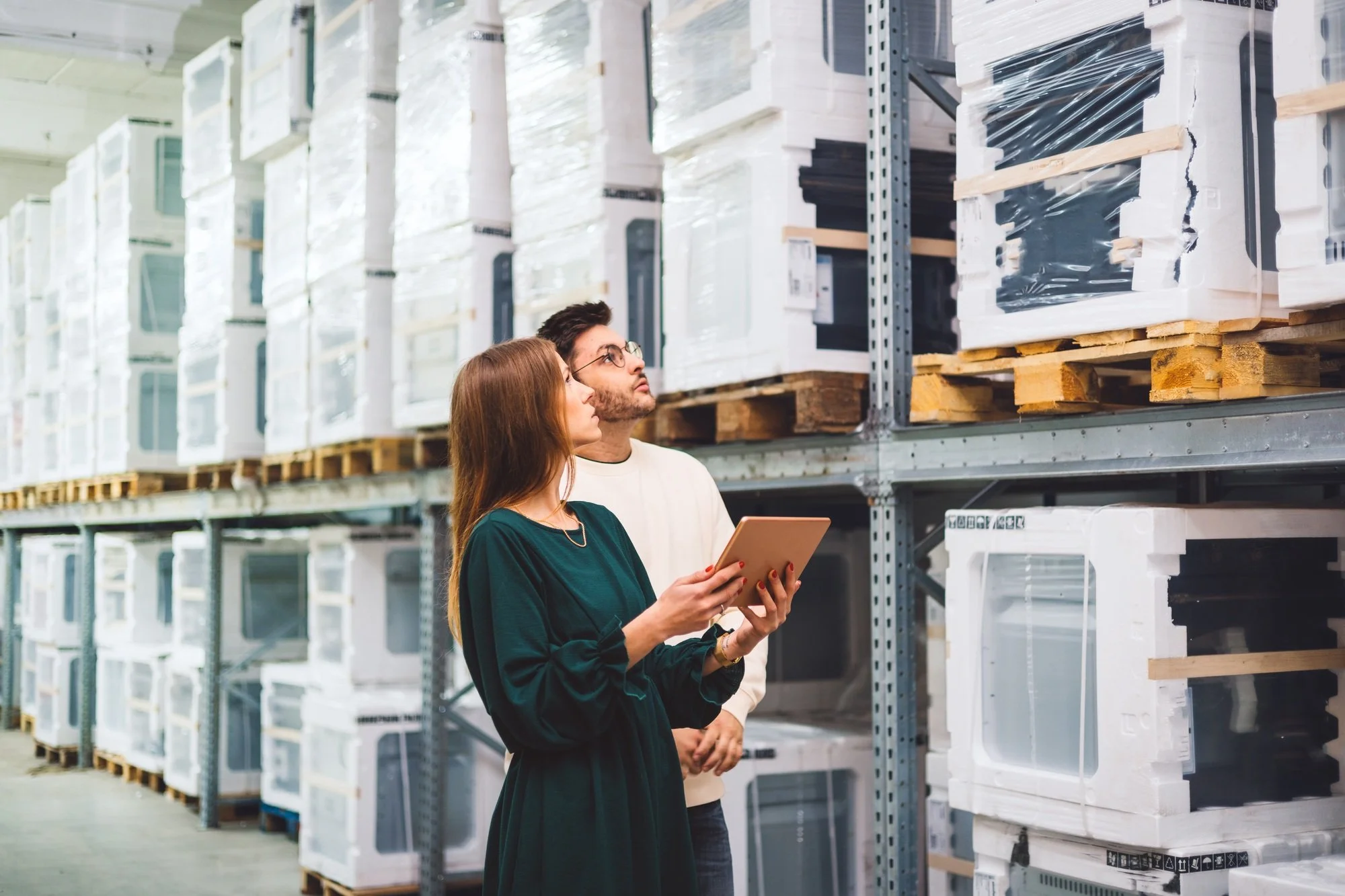 A woman in a dark green dress holding a tablet and a man in glasses and a light-colored shirt looking at stacked white appliances in a warehouse.