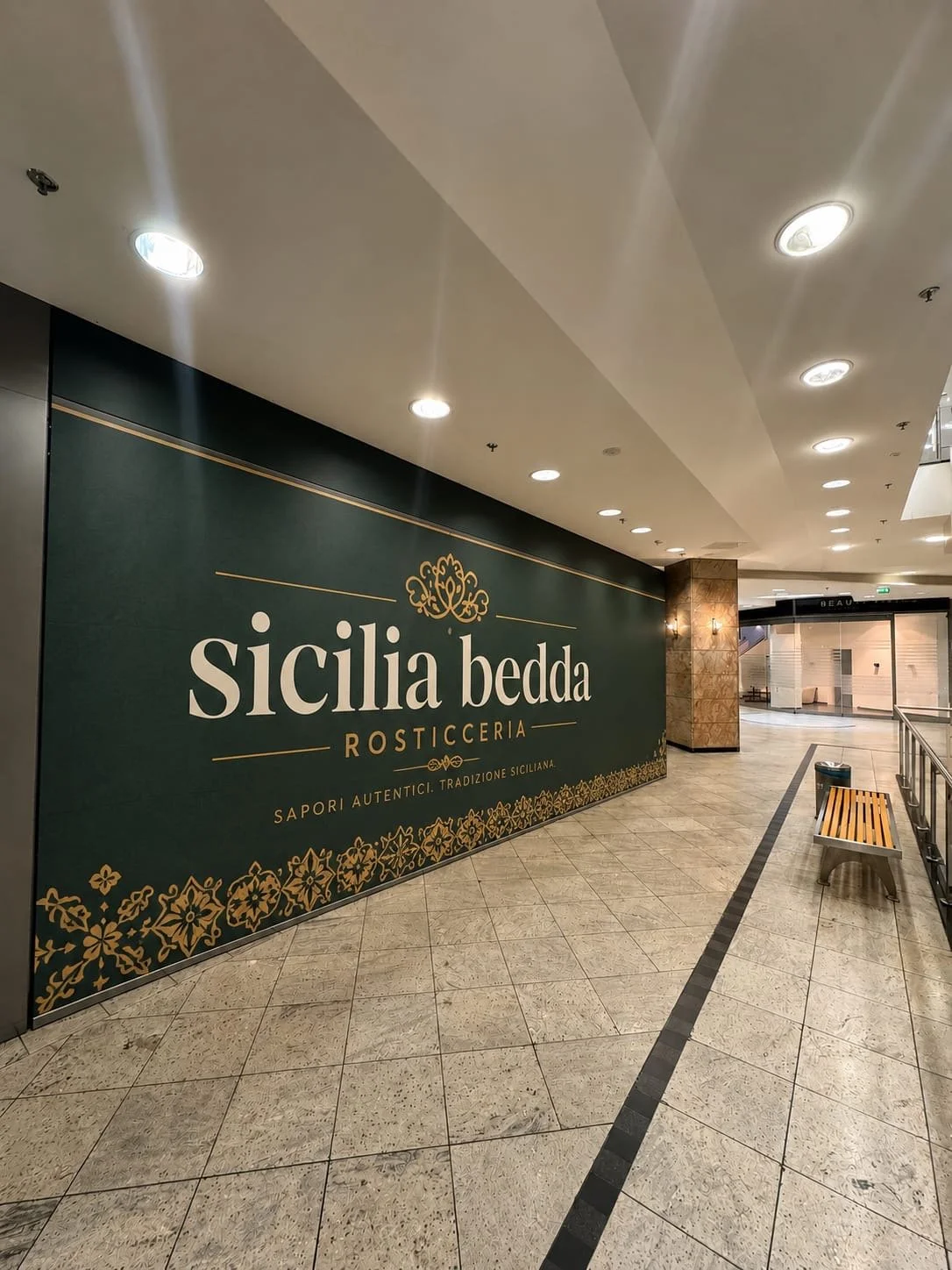 Innenansicht eines Einkaufszentrums mit einem großen dunklen Schild, das 'Sicilia Bedda Rosticceria' mit goldener Verzierung und den Worten 'Sapori autentici. Tradizione Siciliana' an der Wand zeigt. Es gibt eine obere Beleuchtung und einen Sitzbereich mit Bänken.