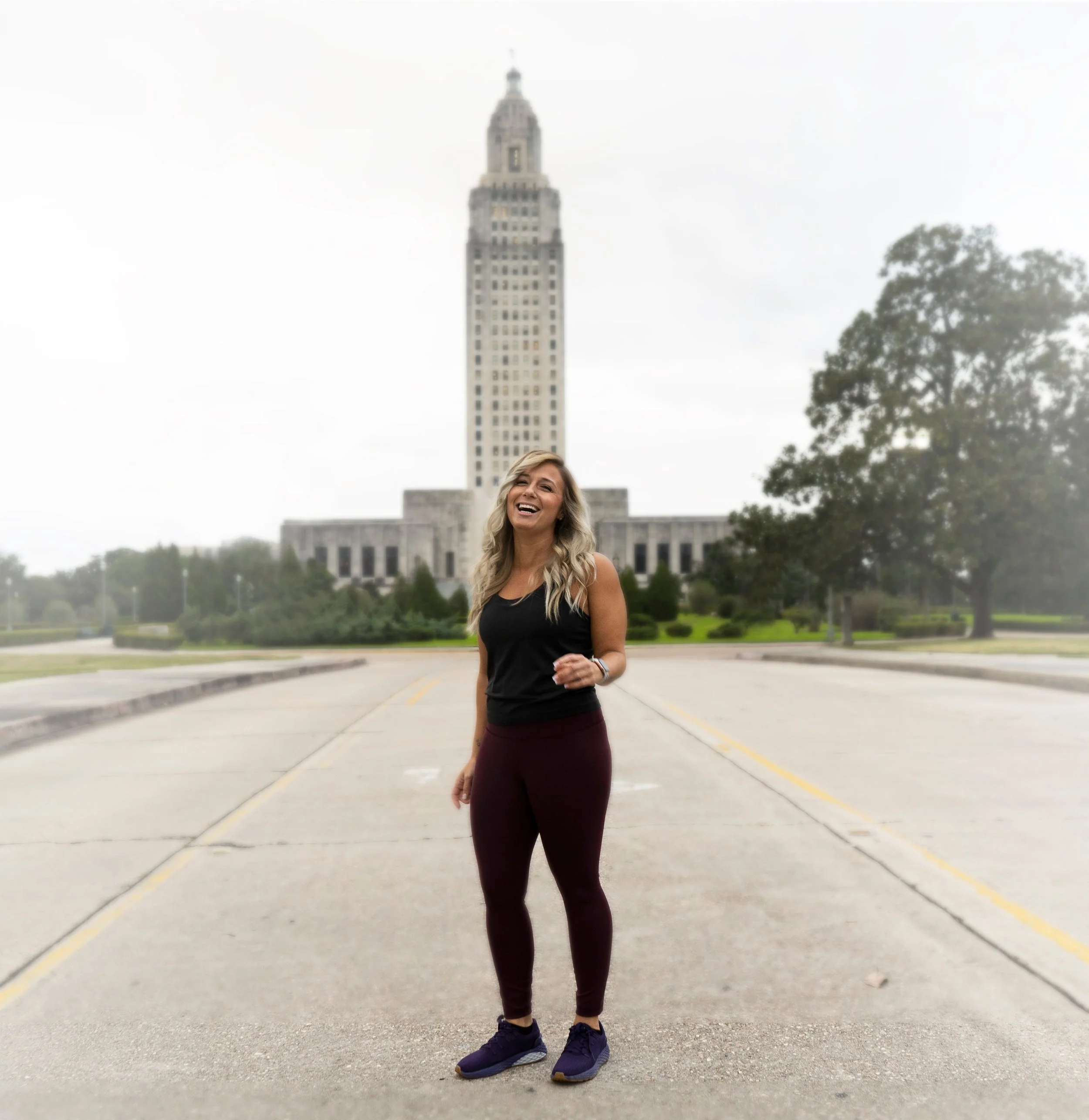 A woman with long blonde hair smiling and running on a city street with the Empire State Building in the background, wearing a black sleeveless top, dark leggings, and running shoes.