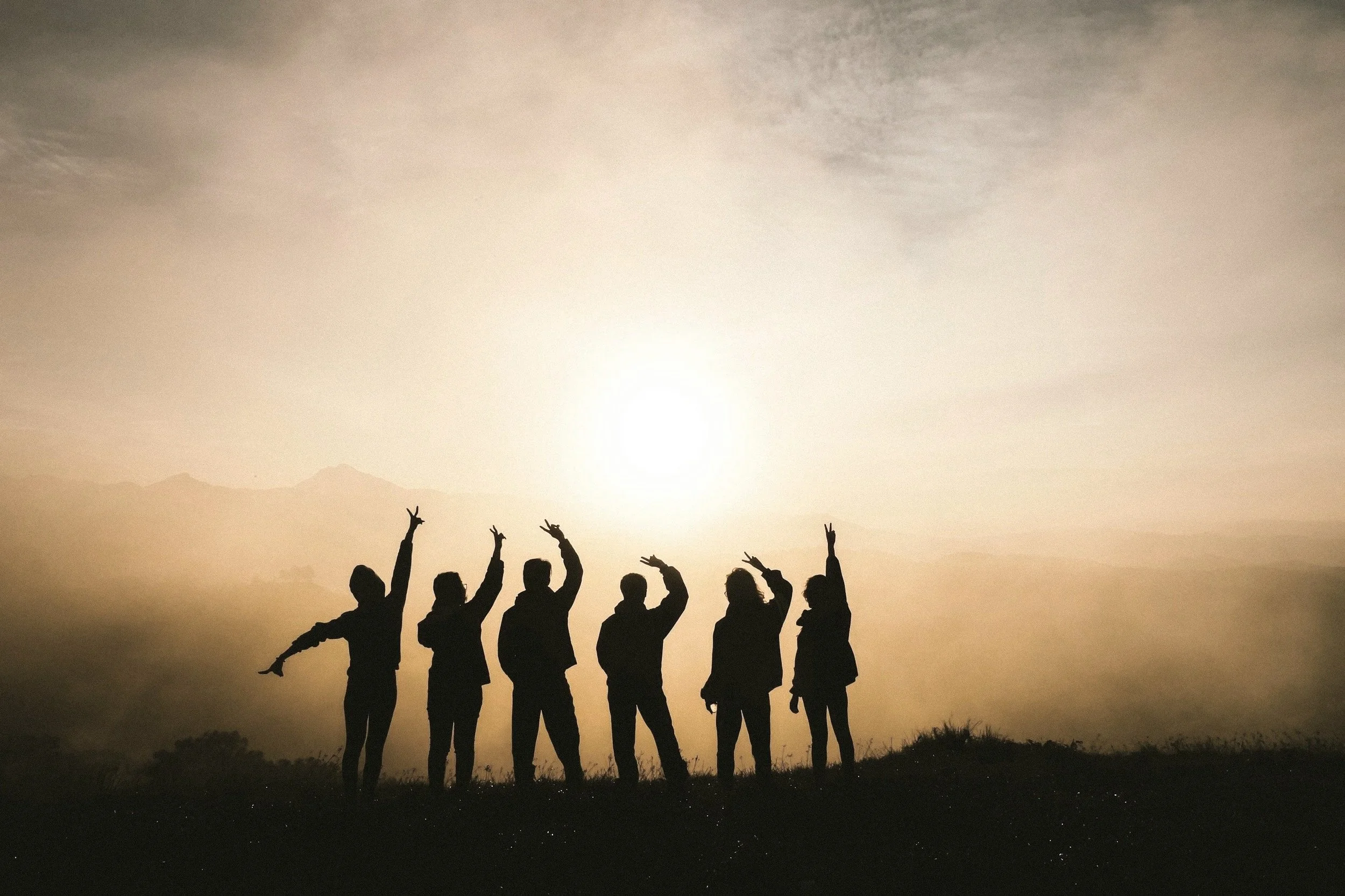 Silhouette di sei persone che fanno il gesto della pace con le mani, sullo sfondo di un tramonto con cielo nuvoloso e montagne all'orizzonte.