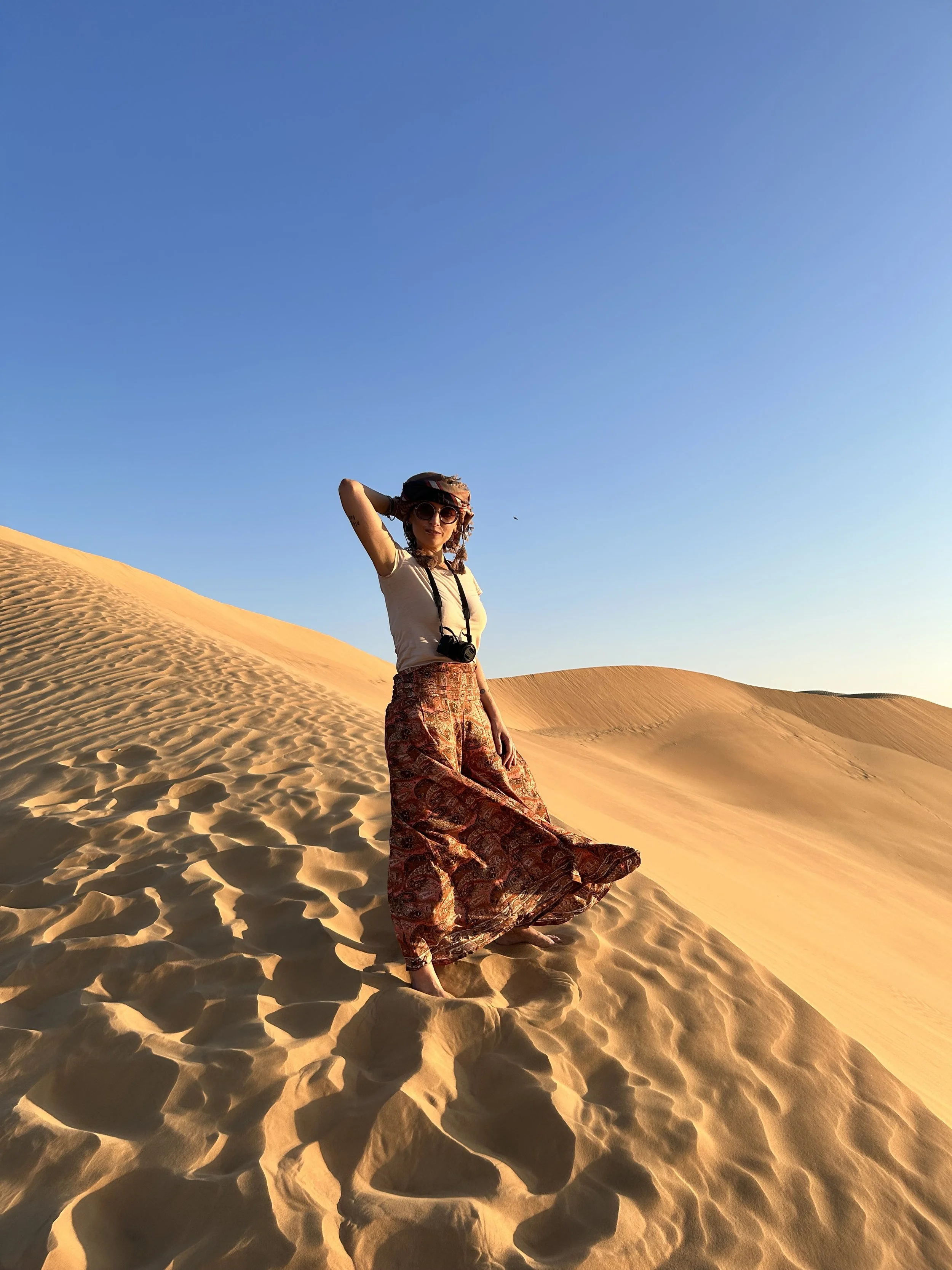 Una donna indossa occhiali da sole, un cappello e abiti ampi, si trova sulle dune di un deserto al tramonto.