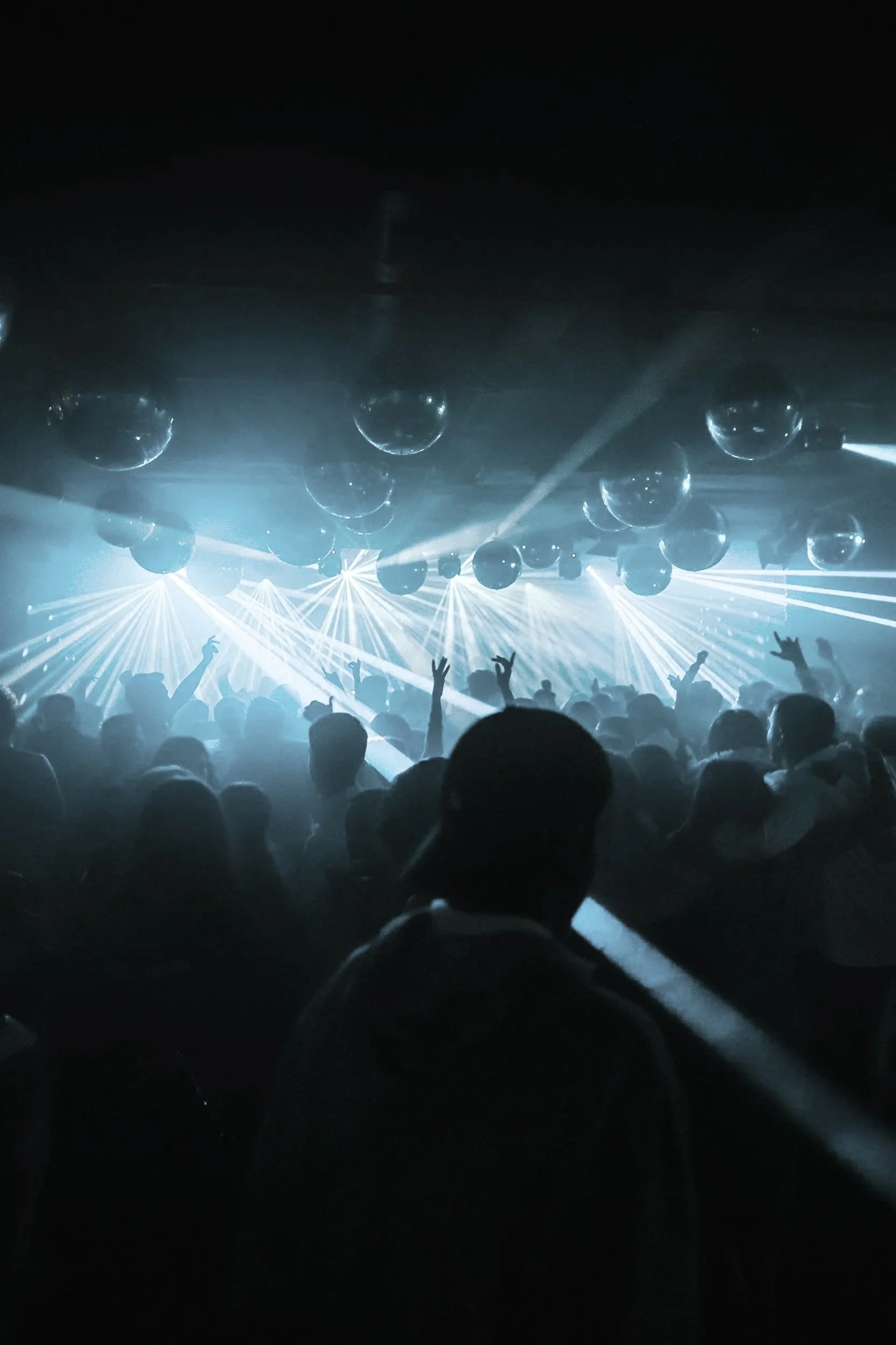 Una scena di un concerto o discoteca con luci soffuse, persone che si divertono e ballano, illuminazione intensa e sfumata, con palloni o decorazioni sospese sopra la folla.