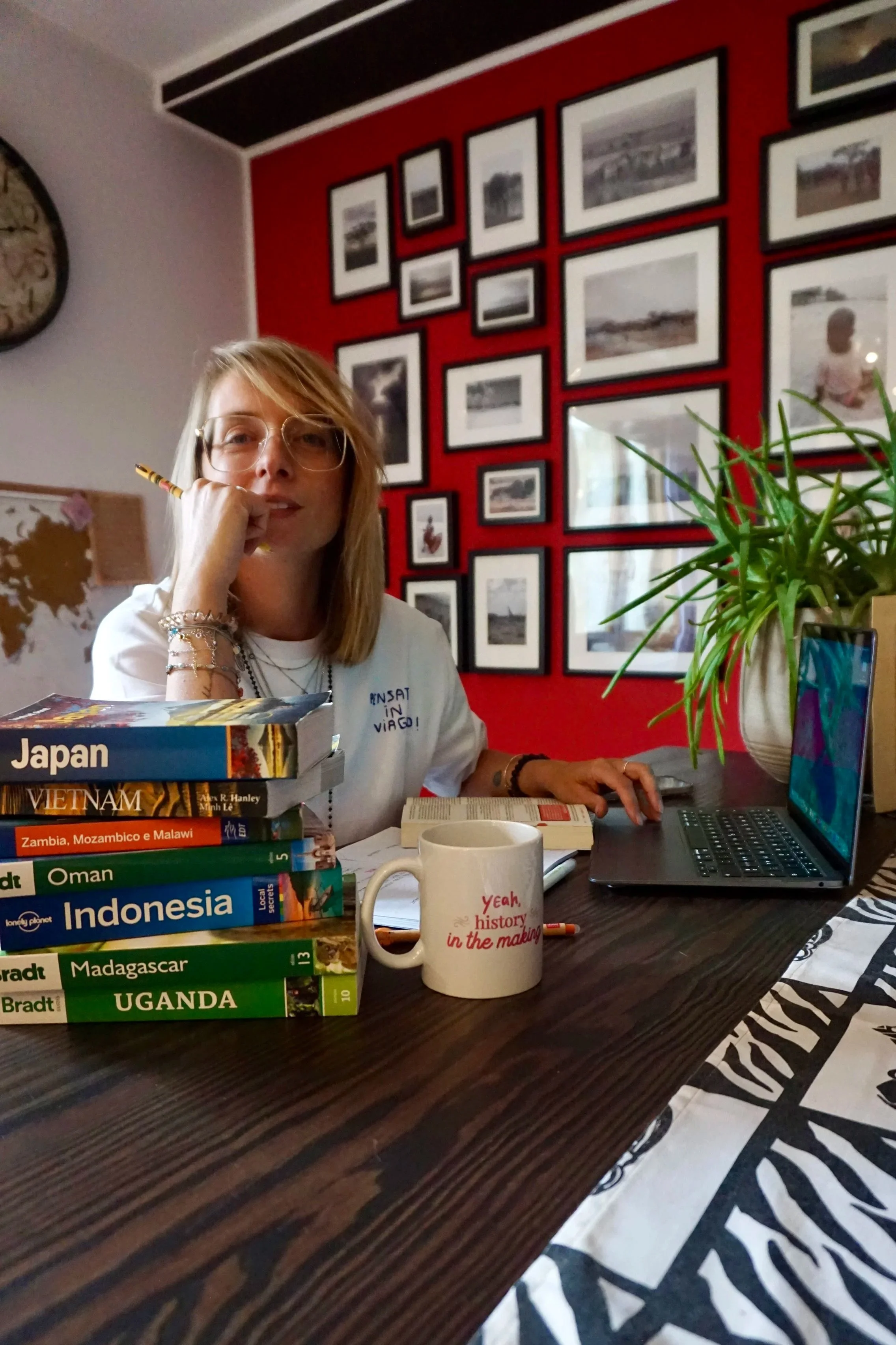 Una donna con occhiali seduta a un tavolo con una pila di libri di viaggio su paesi come Japan, Vietnam, Namibia, Oman, Indonesia, Madagascar e Uganda, una tazza bianca con scritte rosse, un laptop e un mazzo di carte, in una stanza decorata con una parete rossa con molte foto in cornice e un grande orologio sul muro.