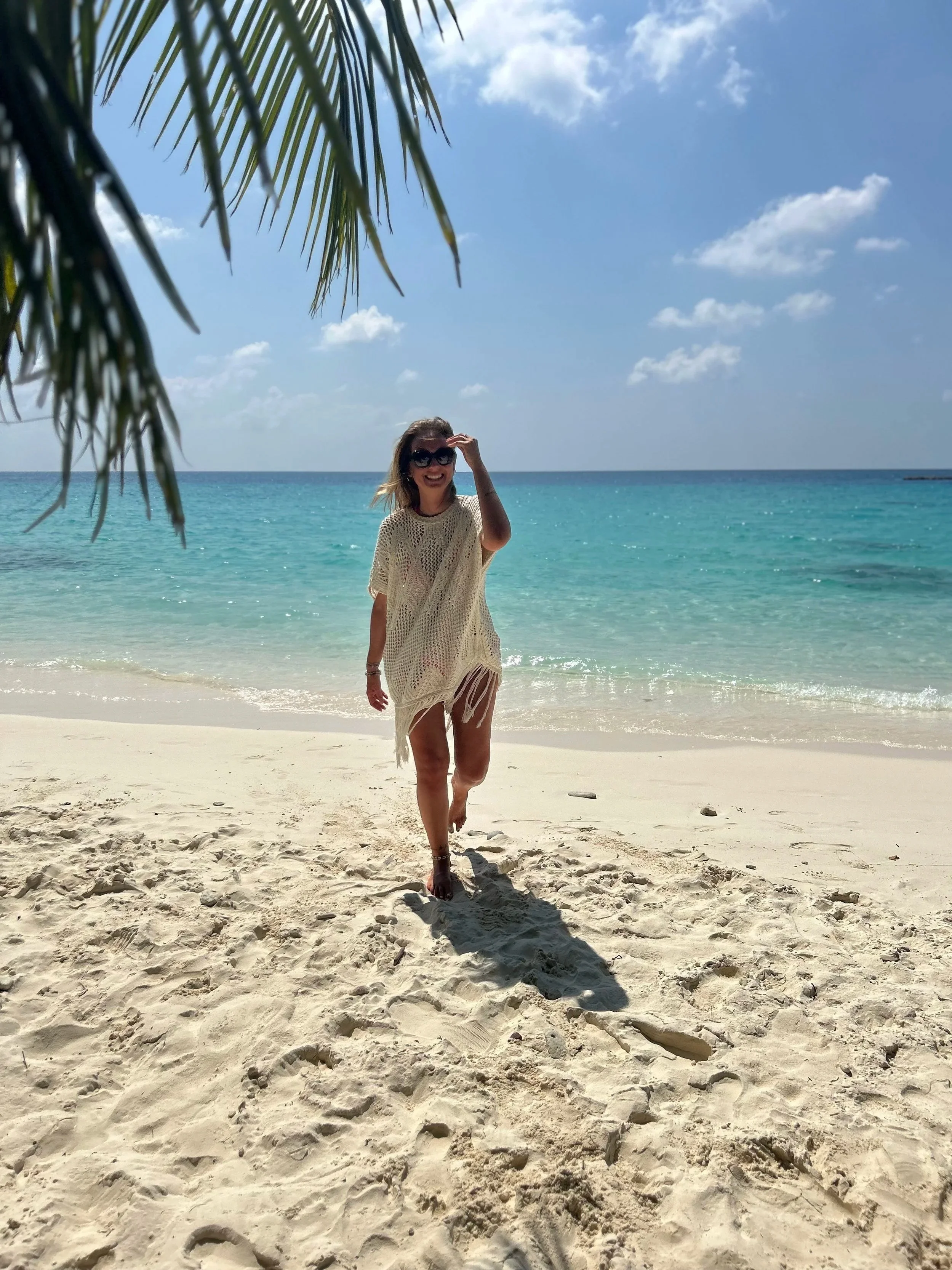Donna sorridente con occhiali da sole, vestita con un maglione color crema, che cammina sulla spiaggia con sabbia chiara e mare turchese sullo sfondo, sotto un cielo azzurro con nuvole sparse.