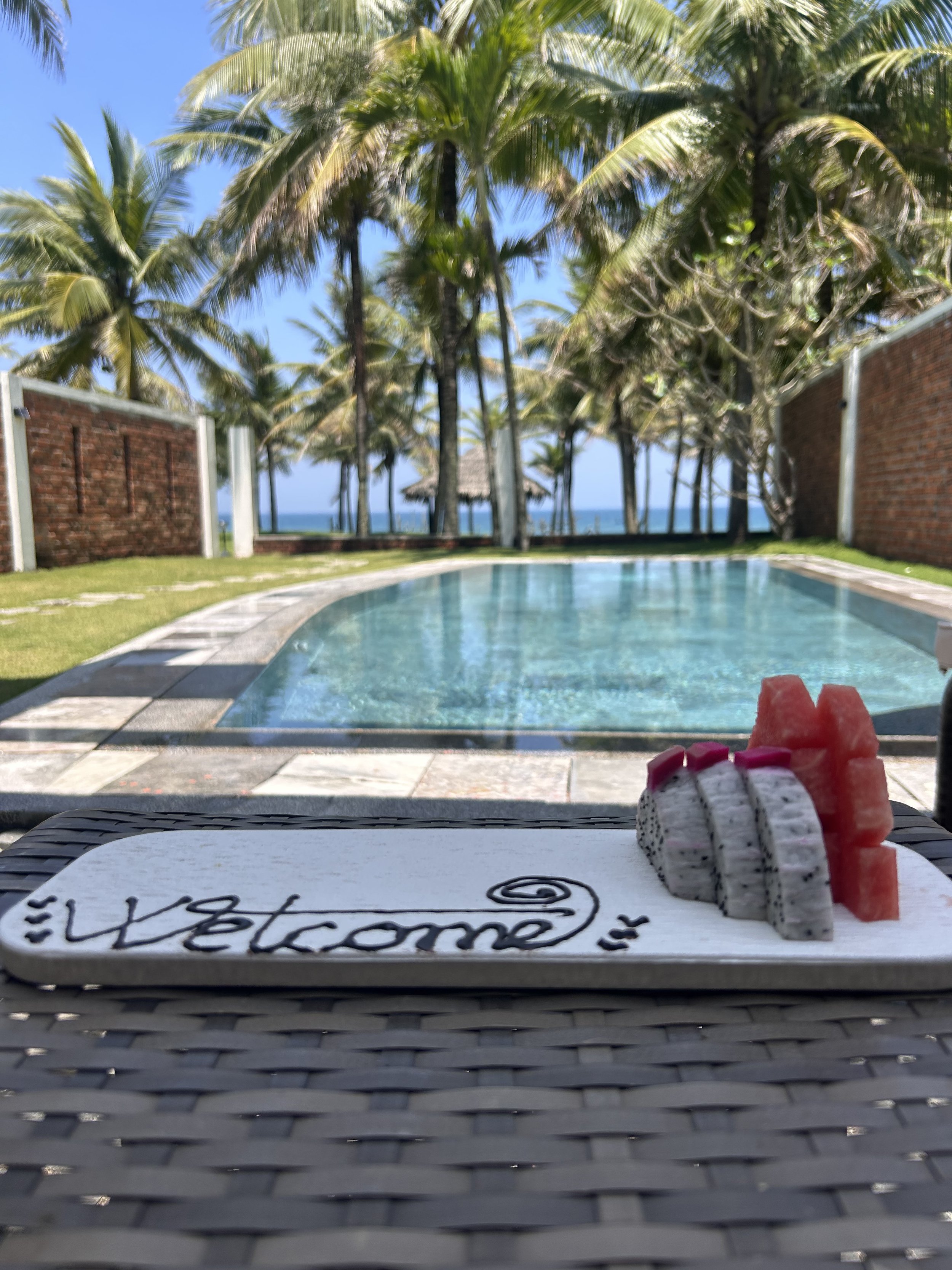 Veduta di una piscina con sfondo di palme, gazebo e vista verso il mare, con una tavola quadrata sulla quale sono posate due decorazioni di sabbia che rappresentano pietre e un biglietto scritto 80° Welcome". In primo piano, un biglietto con scritte in italiano e decorazioni di sabbia.
