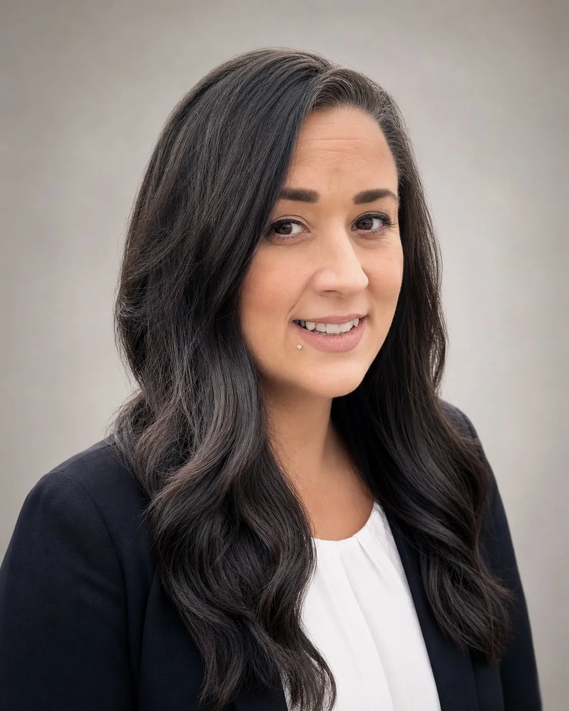 A woman with long, wavy dark hair, wearing a black blazer over a white top, smiling softly with a neutral background.