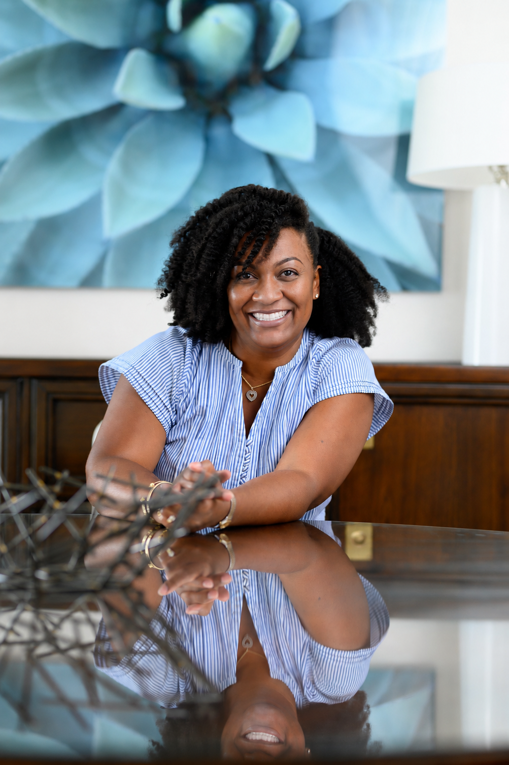 A woman sitting at a reflective wooden table in a modern room with a large blue flower painting behind her, smiling at the camera.