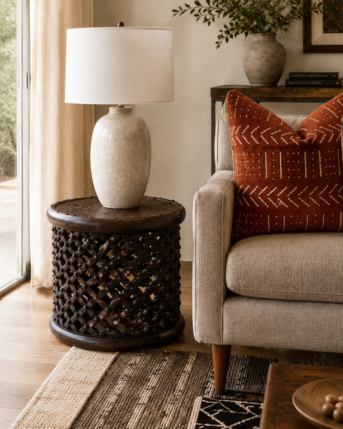 A close-up of a beige sofa with a red patterned throw pillow, next to a side table with a textured ceramic lamp, in a cozy living room with wooden flooring and a rug.