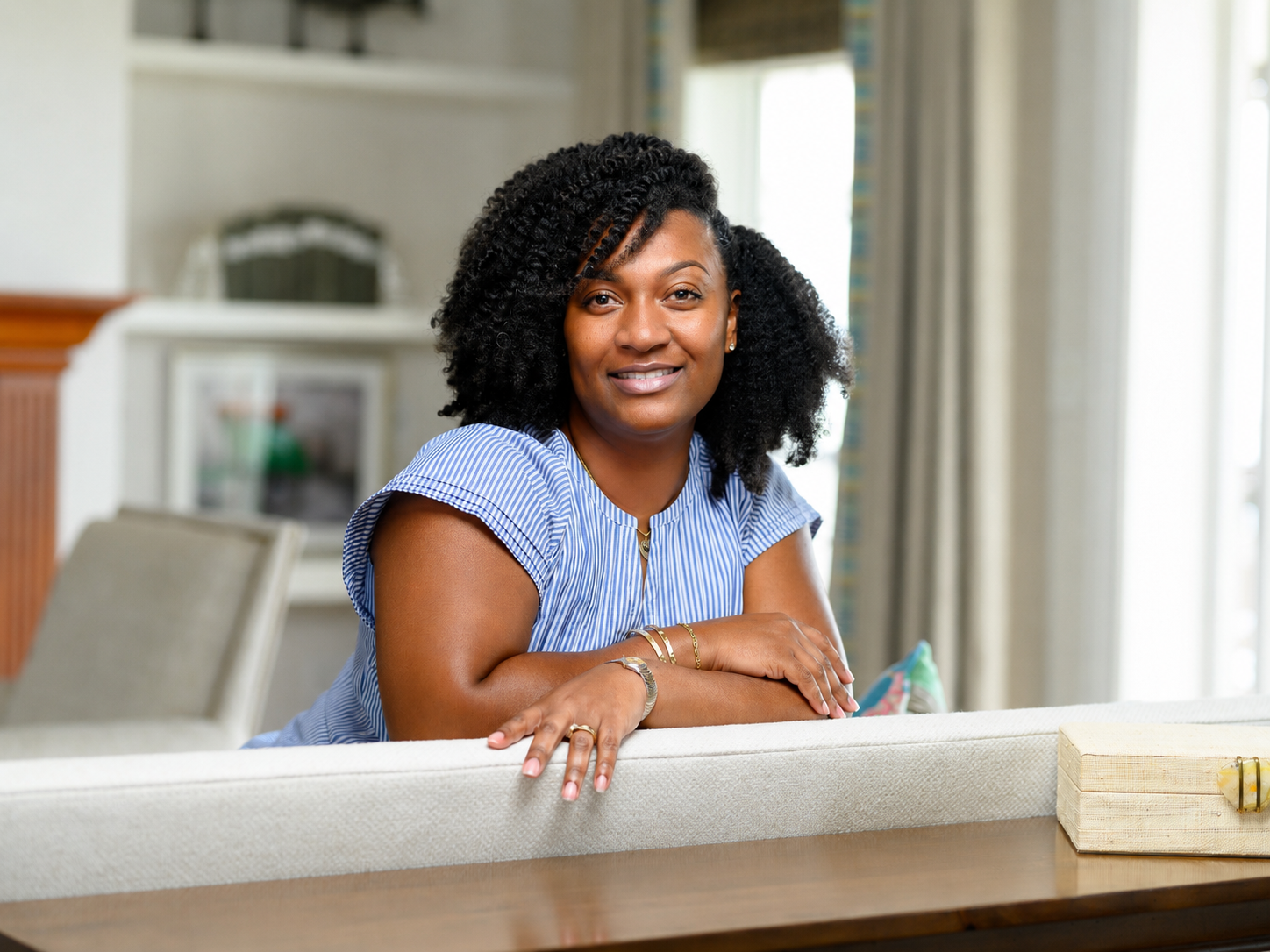Smiling woman with curly black hair sitting at a dining table in a bright room with windows, wearing a blue and white striped top and gold jewelry.