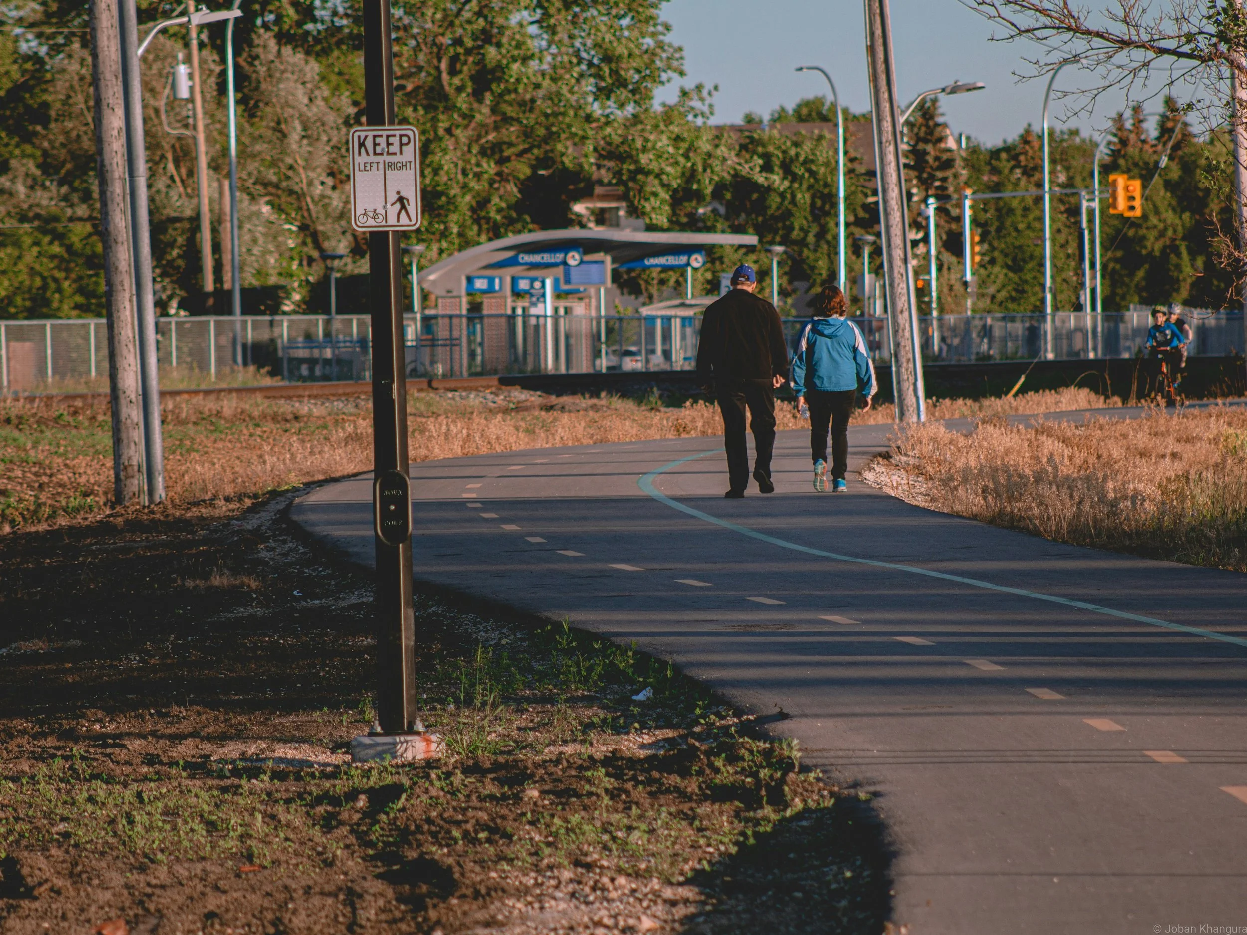 Two people walk hand in hand along a paved bike and pedestrian path in a park, with trees in the background and a sign indicating keeping left or right for bikes and pedestrians.