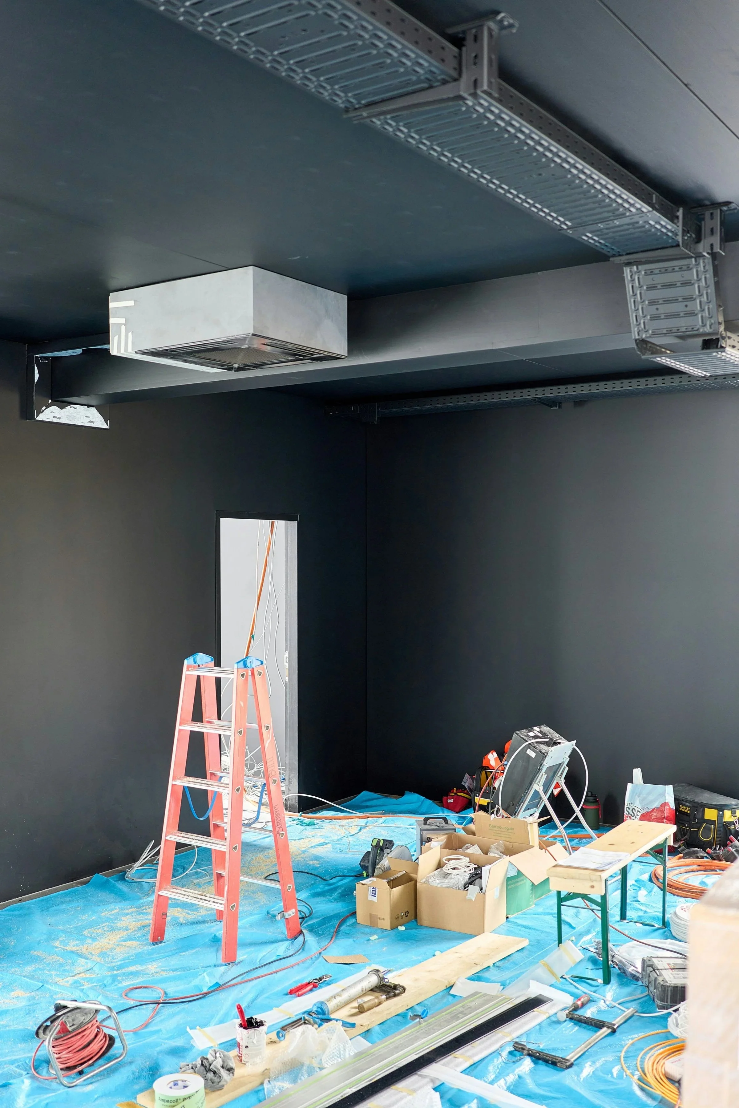 Construction site indoor room with black walls and ceiling, electrical and HVAC work in progress, tools, wires, and equipment scattered on blue protective floor covering, a red ladder, and an opening in the wall leading to another room.