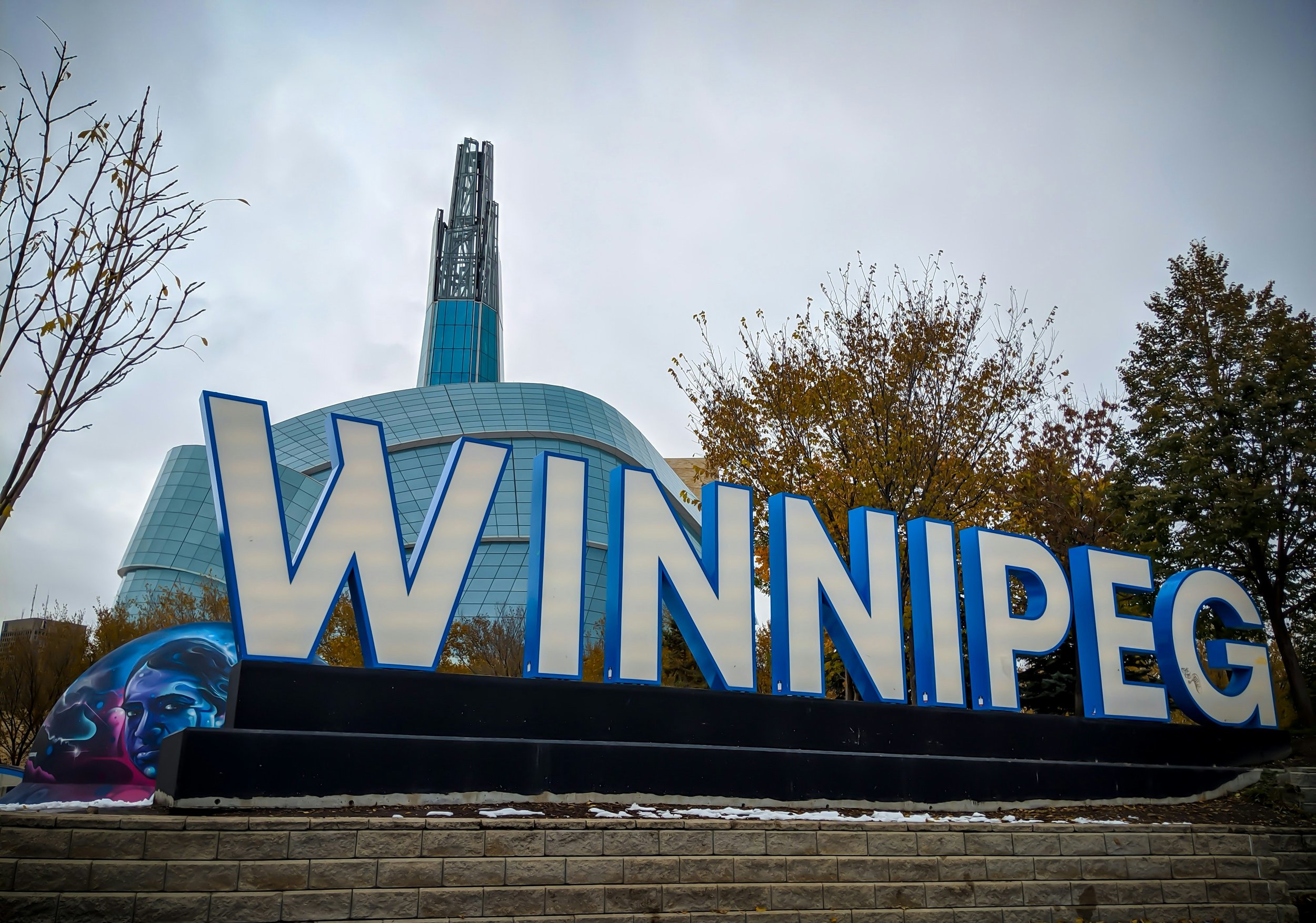 A sign with the word 'WONDER' in large blue letters in front of a modern, curved skyscraper with a spire, trees with autumn leaves, and an overcast sky.