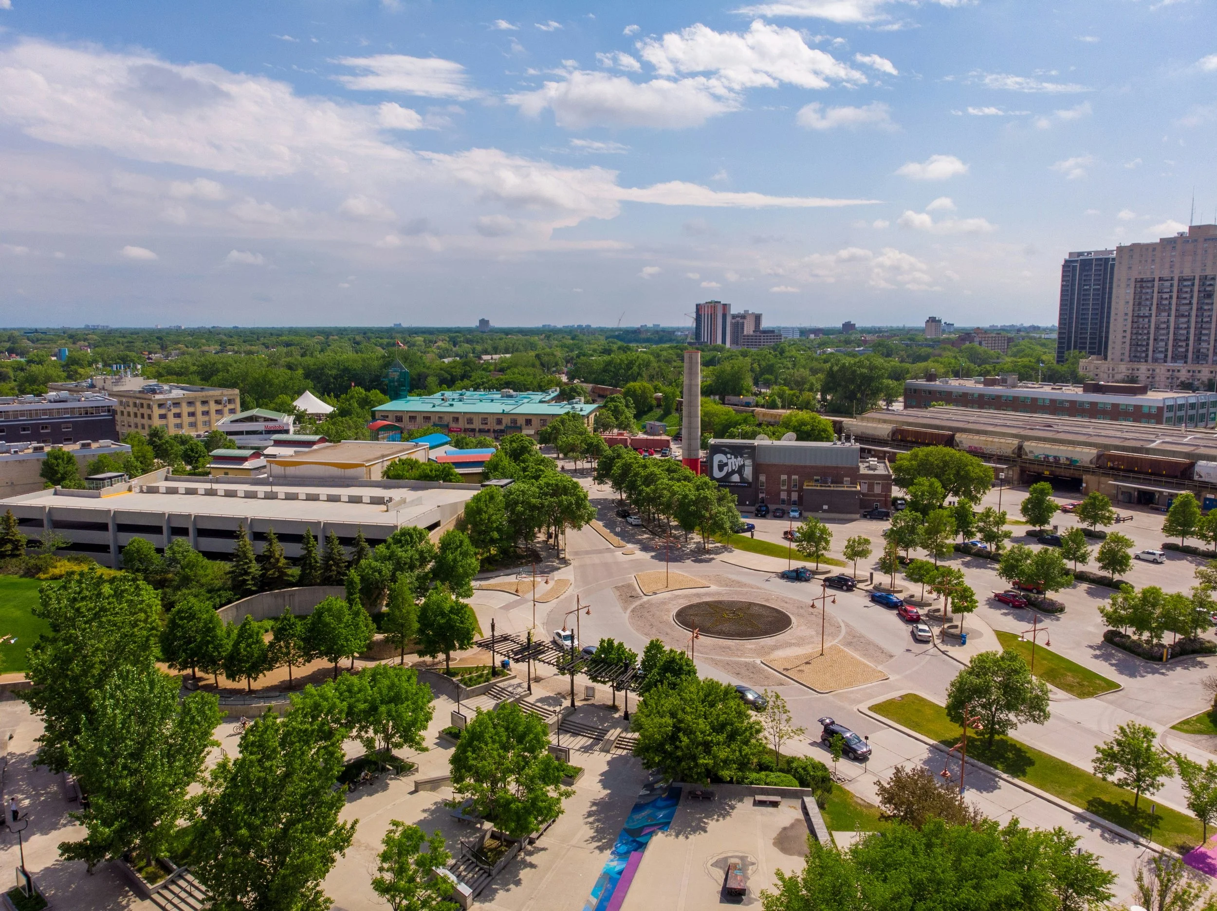 A cityscape view showing a mix of tall buildings, shorter structures, and lush green trees under a partly cloudy sky.