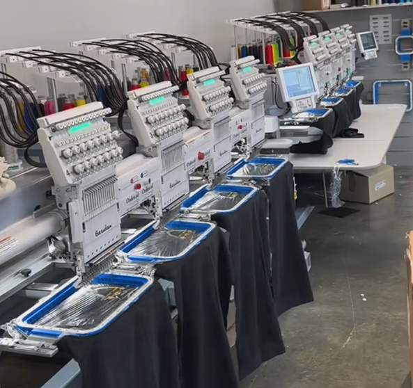 Multiple industrial embroidery machines set up on a long table with black skirts and blue trays underneath, in a workshop or factory environment.