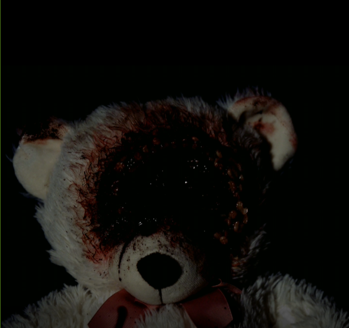 A stuffed teddy bear with bloodstains on its face and eyes, against a dark background.