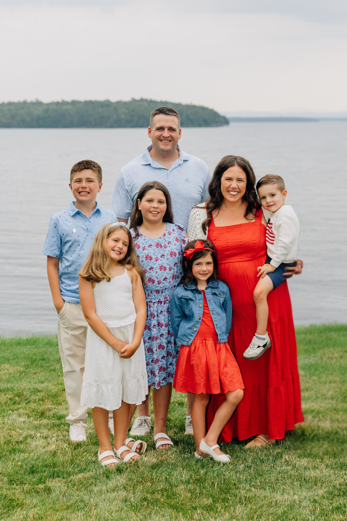 A family of eight standing on grass near a body of water with a forested island in the background. The family includes three adult women, one adult man, and four children.