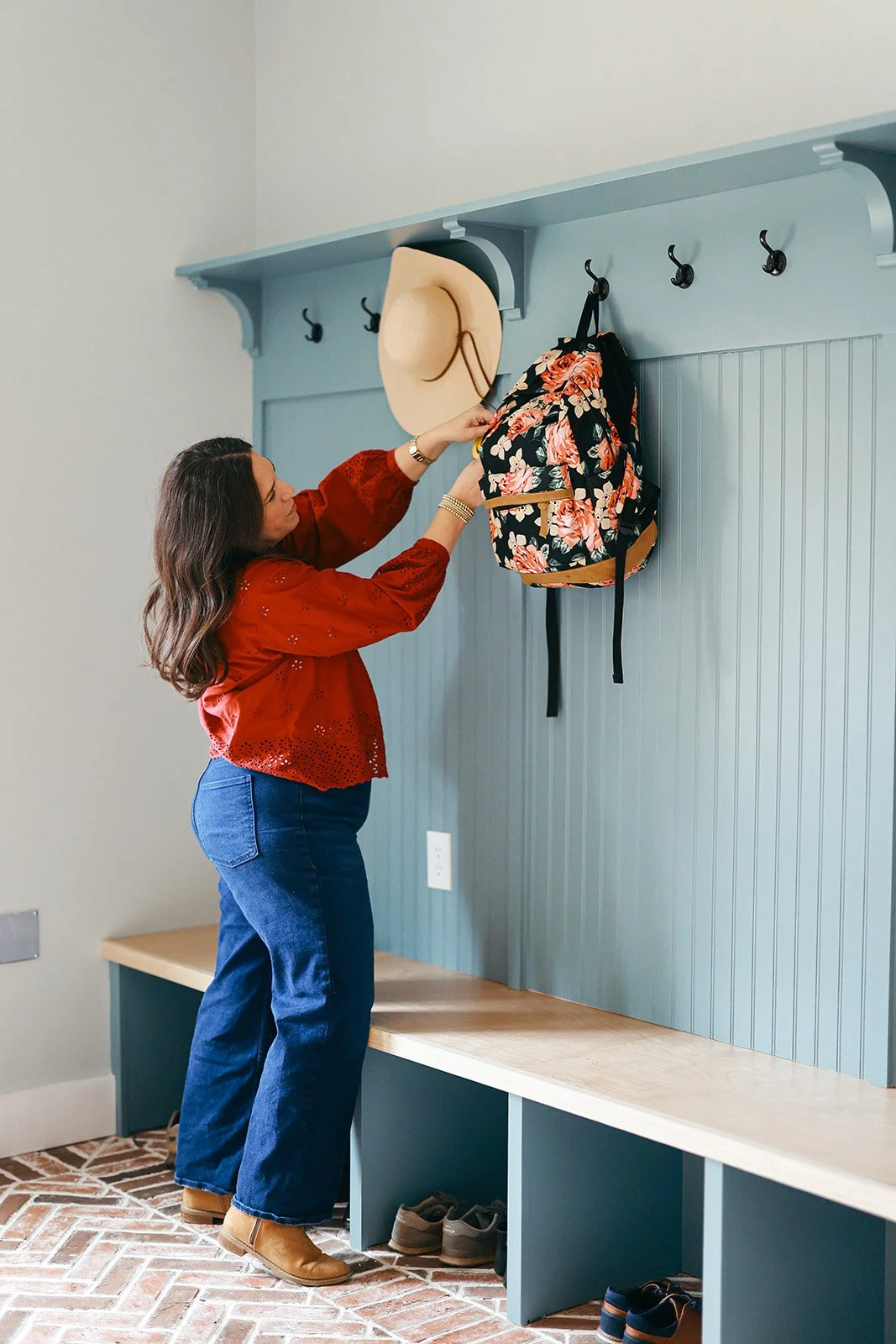A woman in a red blouse and blue jeans hangs a floral backpack on a hook in a blue wall-mounted coat rack above a wooden bench.