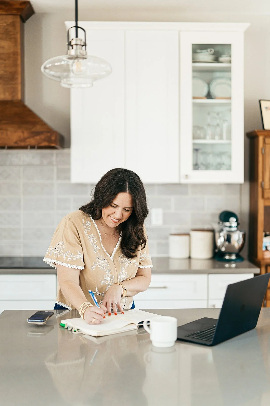 Woman writing in a notebook at a kitchen island, with a laptop, mug, and phone nearby.