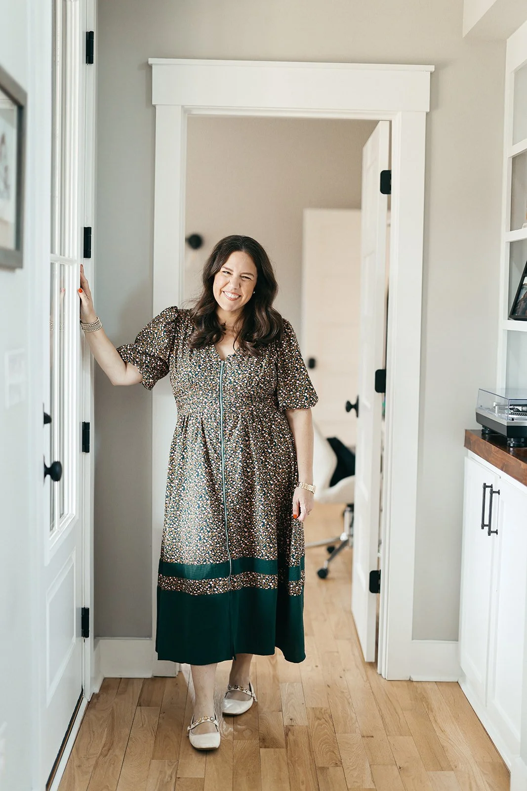 Woman in a floral dress smiling and standing in a doorway, with a hardwood floor and part of a room visible behind her.