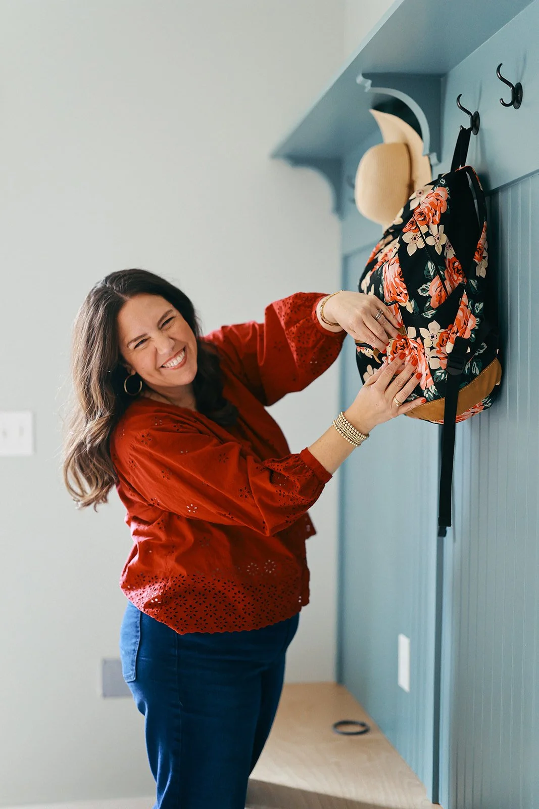 Woman with long brown hair smiling and hanging a floral backpack on a hook on a blue wall.
