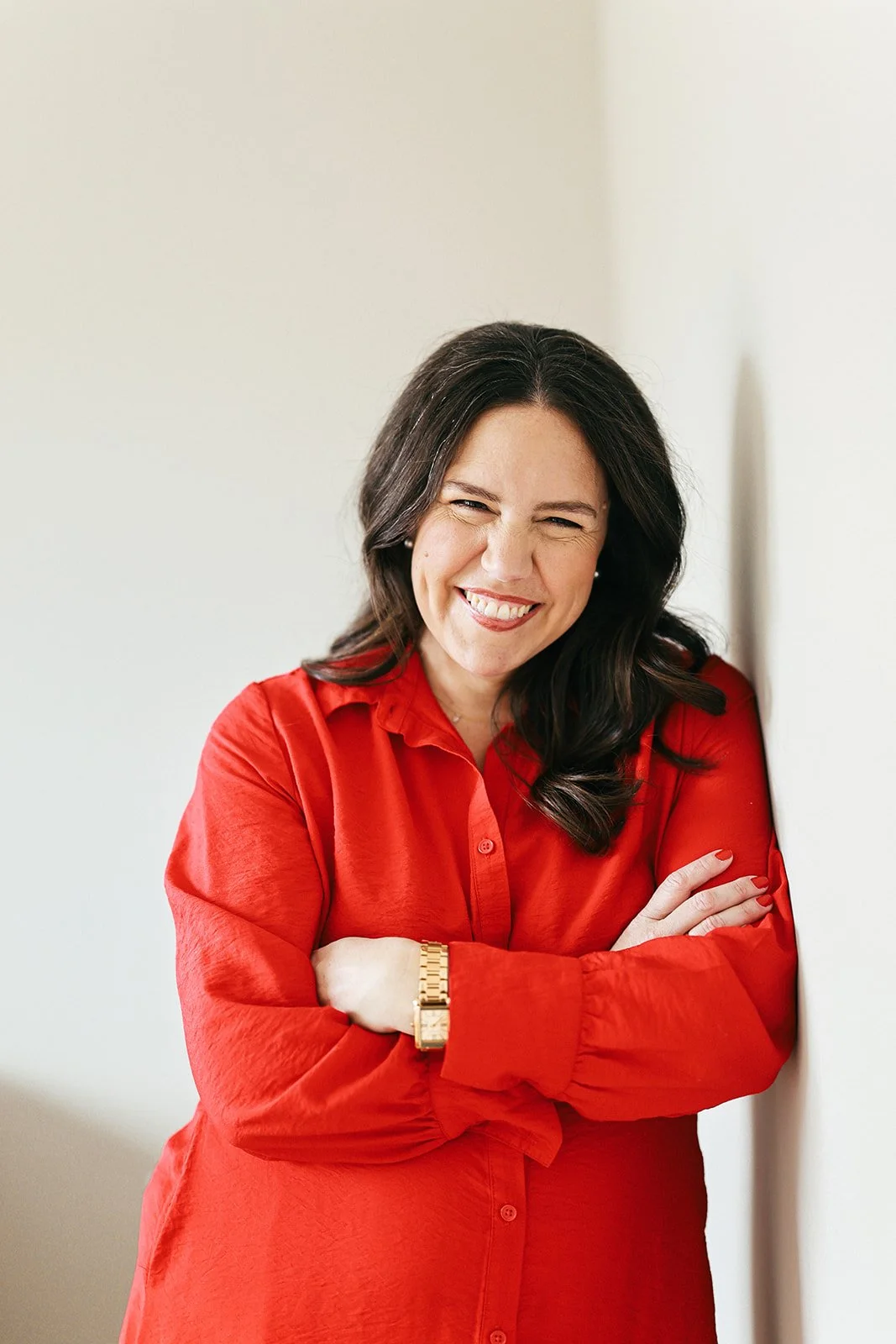 A woman with dark hair smiling and wearing a red blouse, leaning against a wall.