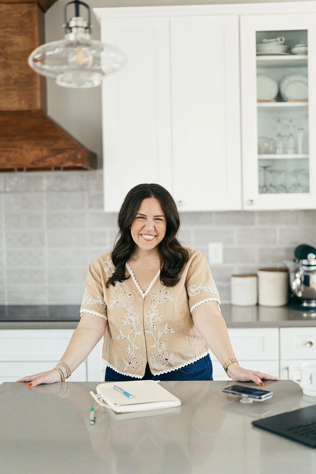 A woman with dark hair smiling while leaning on a kitchen countertop, with a notebook, pen, and phone in front of her.