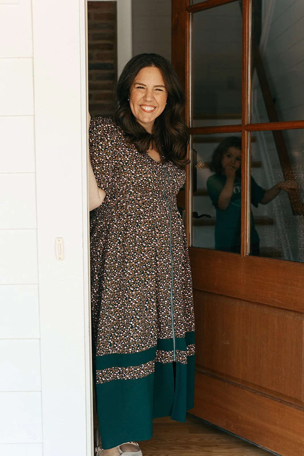 A woman with dark hair smiling and peeking out from behind a white wall, standing by a wooden door with glass panels. A young girl is visible in the reflection in the glass, standing inside the house.