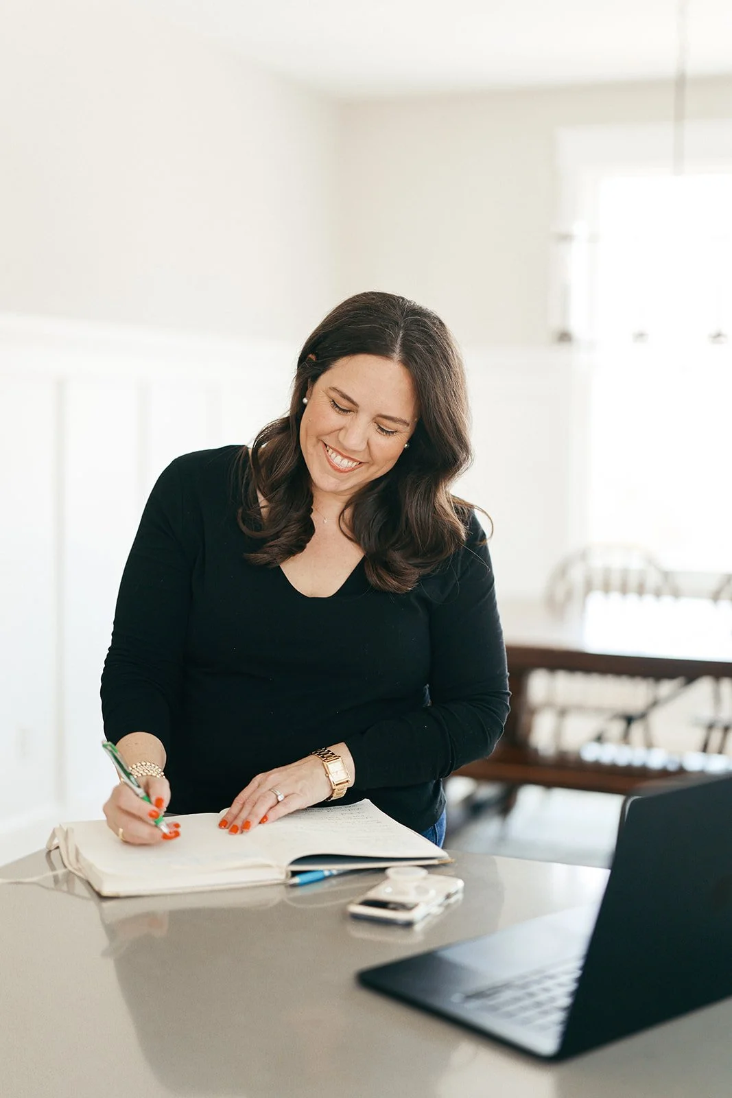 A woman with dark hair smiling while writing in a notebook on a kitchen table, with a laptop and a smartphone nearby.
