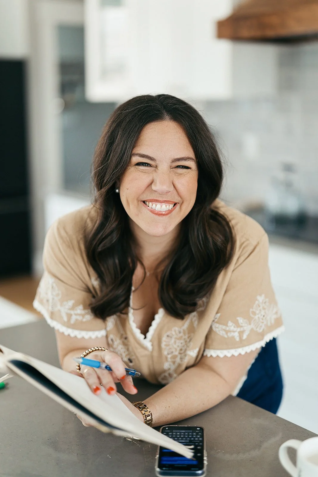 A woman with dark hair and a beige embroidered top smiling and leaning over a table with a notebook, pen, and phone.