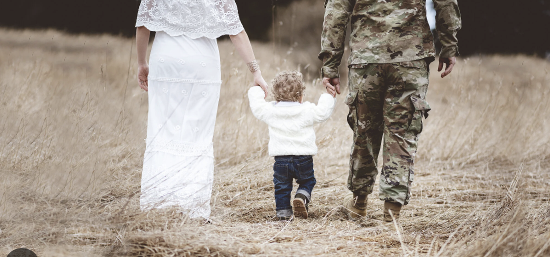 A toddler walking between two adults, one in a white dress and the other in military camouflage, holding his hands in a grassy field.
