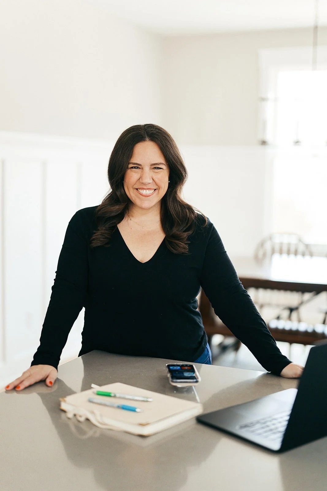 Woman with dark hair smiling, standing at kitchen counter with papers, pens, smartphone, and laptop.