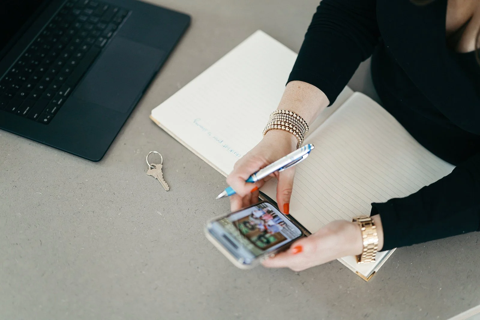 Woman with red painted nails holding a smartphone, sitting at a desk with notebooks, a keyboard, and a key, taking a photo of her phone's screen.